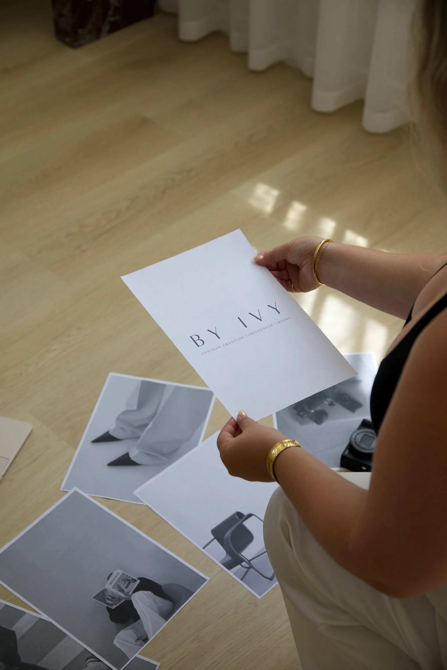A person is holding a white paper with the words "BY IVY" and some additional text, with other printed photos of clothing and chairs on a wooden table.