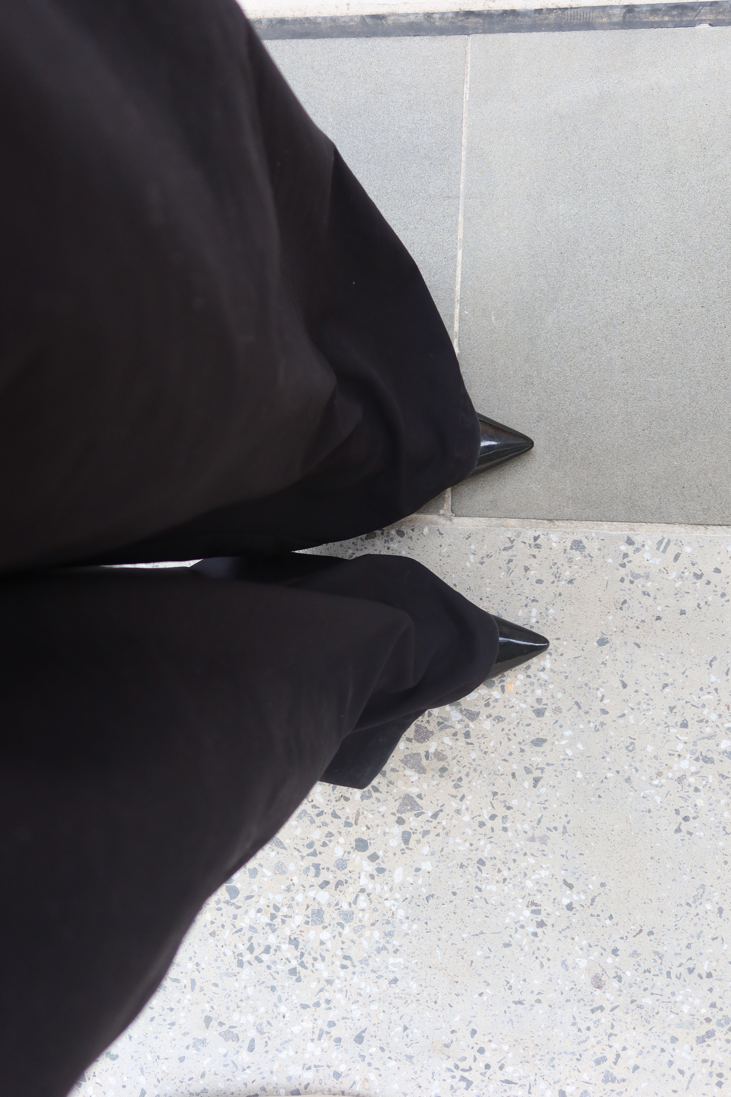 A person wearing black pants and black high-heeled shoes standing on a grey tiled floor.