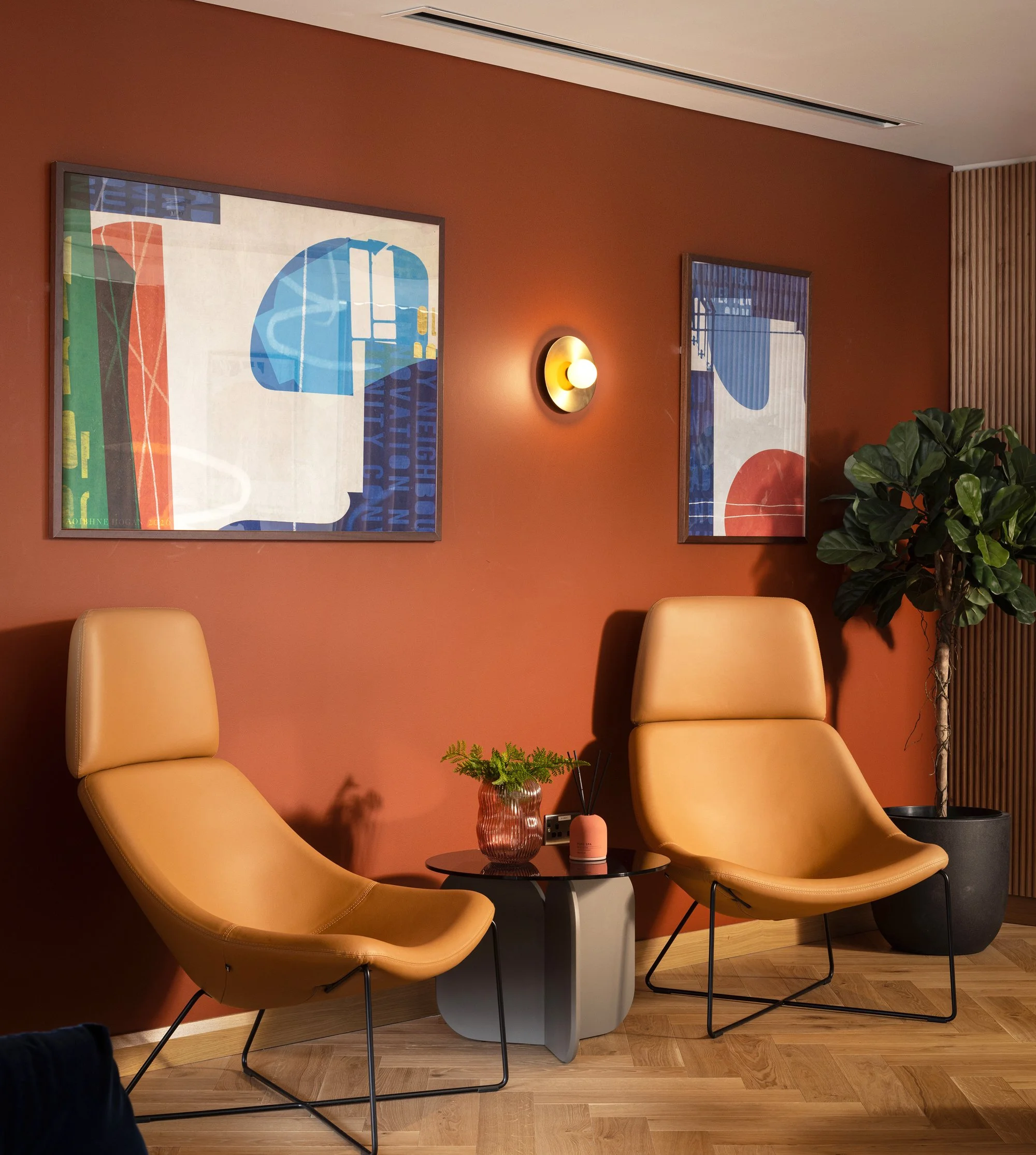 A cozy seating area with four beige chairs arranged around a small round black table on a wooden floor, against a warm terracotta-colored wall decorated with two framed abstract art pieces and a modern wall sconce.