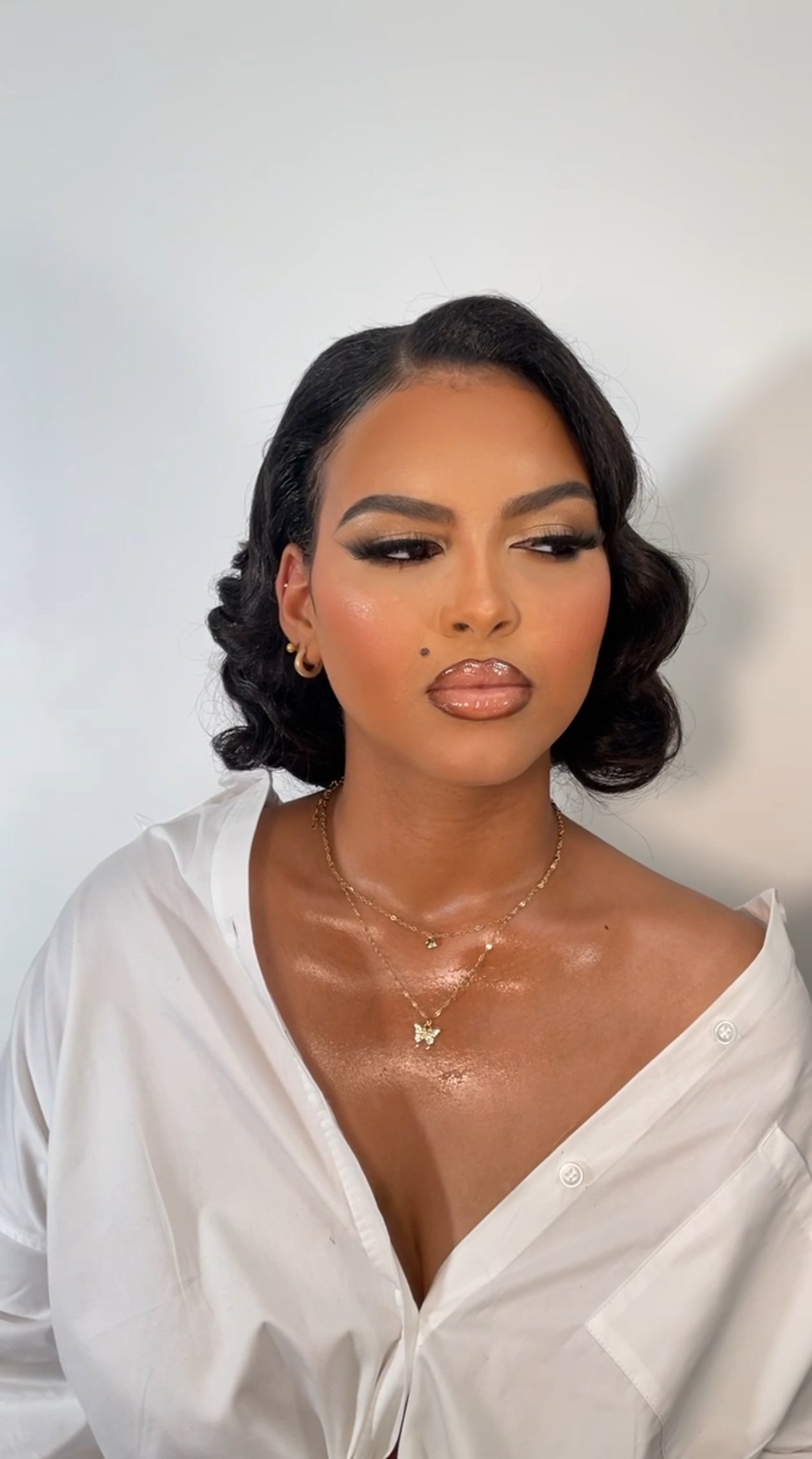 Person with elegant makeup, glossy lips, wearing a white shirt and layered gold necklaces, with short curly hair.