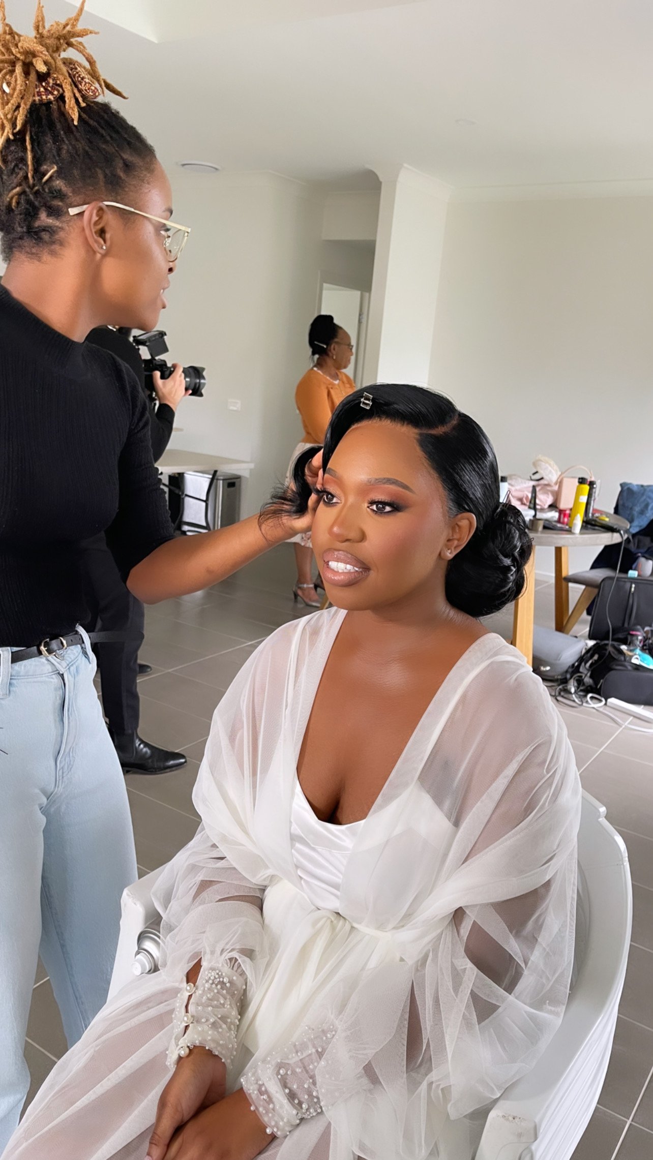 A woman sitting in a white chair wearing a sheer white robe with pearl detailing, receiving makeup and hair styling in a bright room. Another person with dreadlocks and glasses is assisting her. In the background, a person in an orange top and photographer are visible, with styling tools and products on a table.