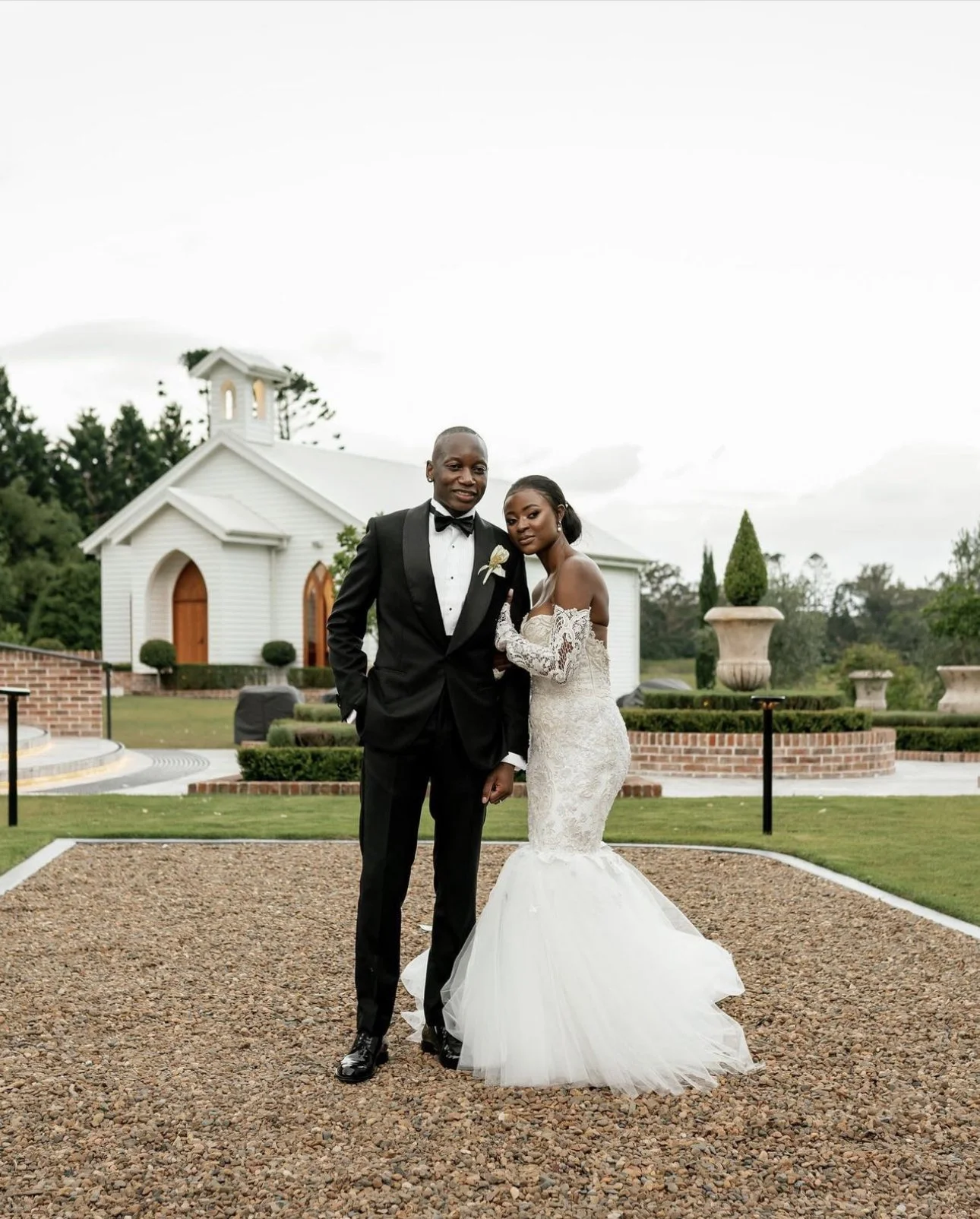 A bride and groom pose in formal attire in front of a small white chapel on their wedding day. The bride wears an off-the-shoulder lace gown, and the groom is in a black tuxedo with a bow tie. The setting includes a well-manicured garden.