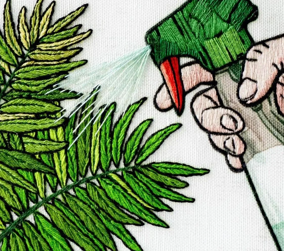 Close-up of embroidered artwork showing a hand spraying water from a green spray bottle onto green fern leaves.