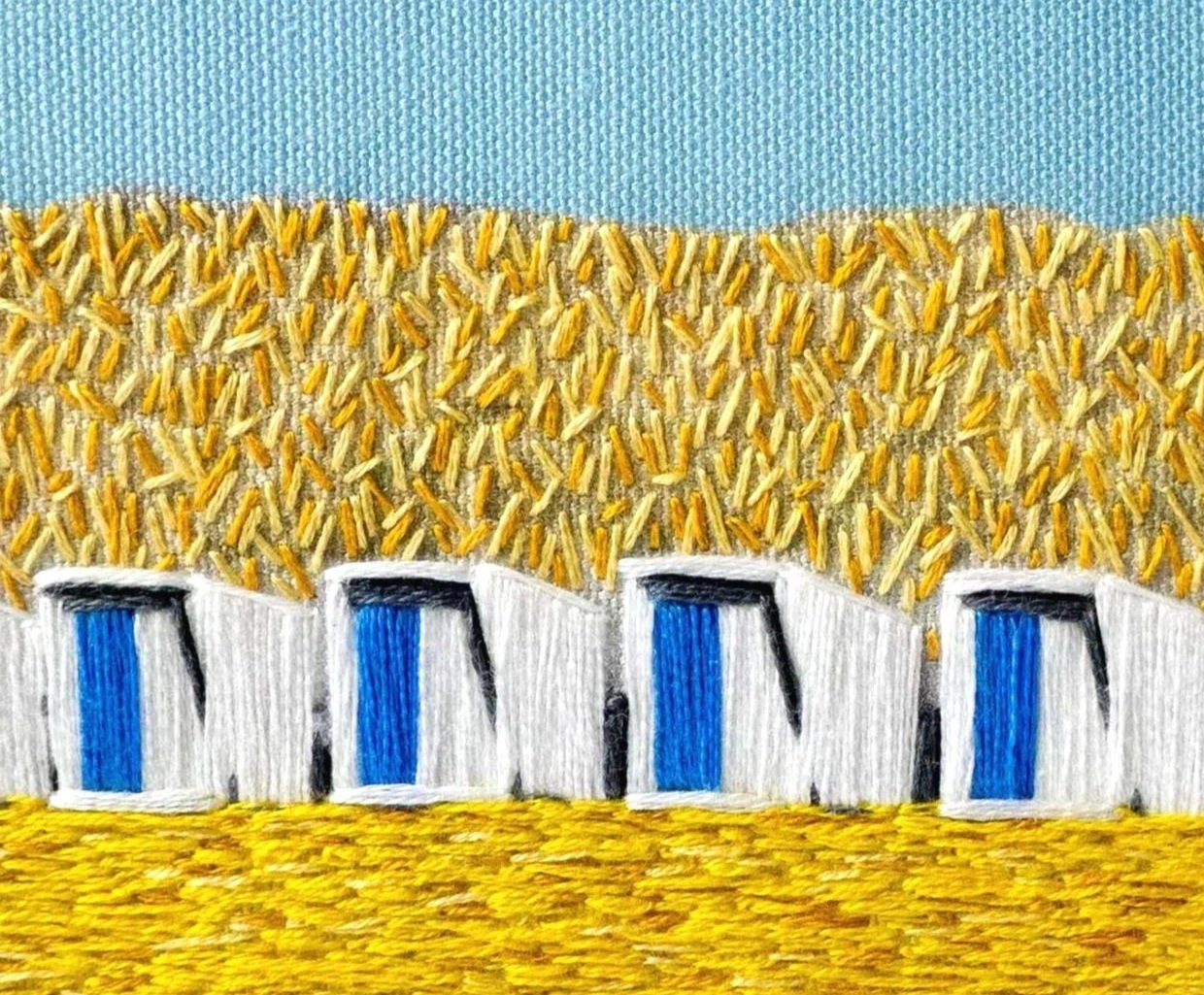 Close-up embroidery of a landscape with blue sky, yellow grass, and beach cabins.