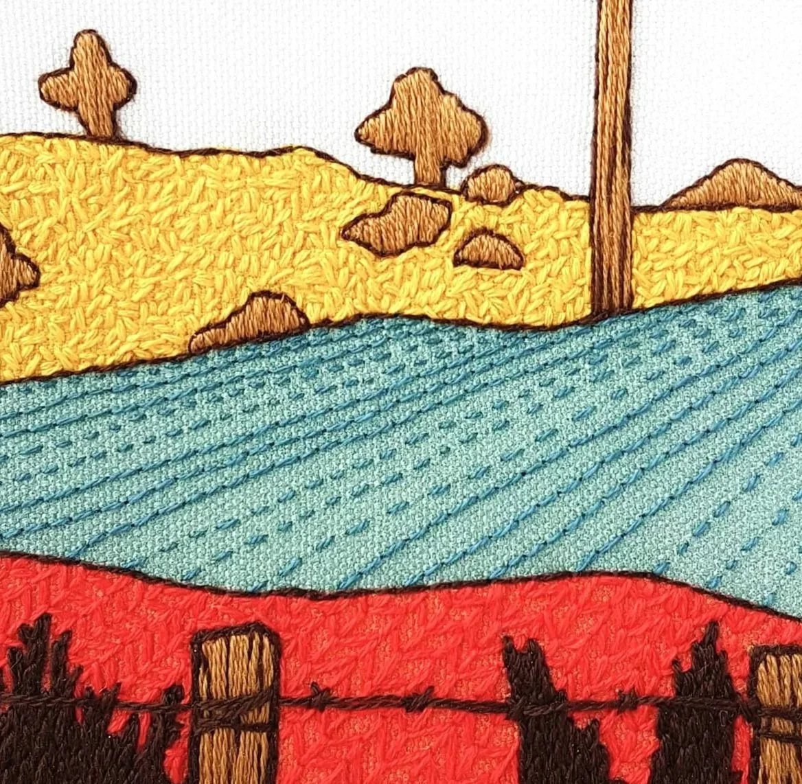 Embroidery of a landscape with power lines, trees, hills, and a fence, featuring yellow, blue, and red sections.