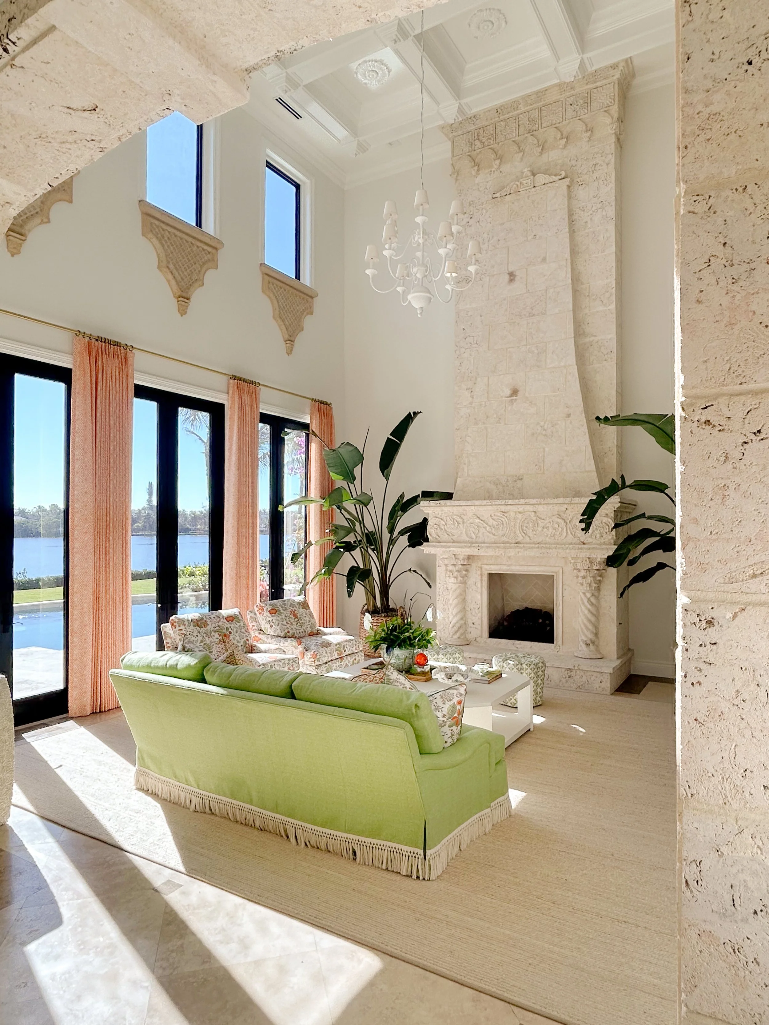 Bright living room with large windows, a white ornate fireplace, a chandelier, potted plants, and pastel furniture, overlooking a scenic water view.