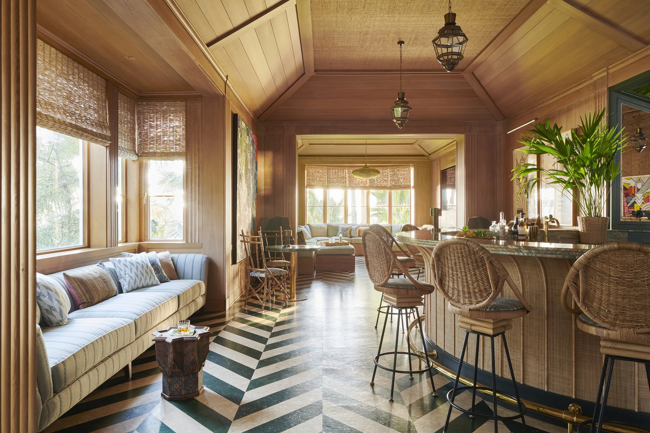 Interior view of a cozy living room with wooden walls and ceiling, large windows with bamboo shades, a striped sofa with multiple pillows, a wooden side table, a bar counter with wicker bar stools, green plants, and hanging lantern-style lights.