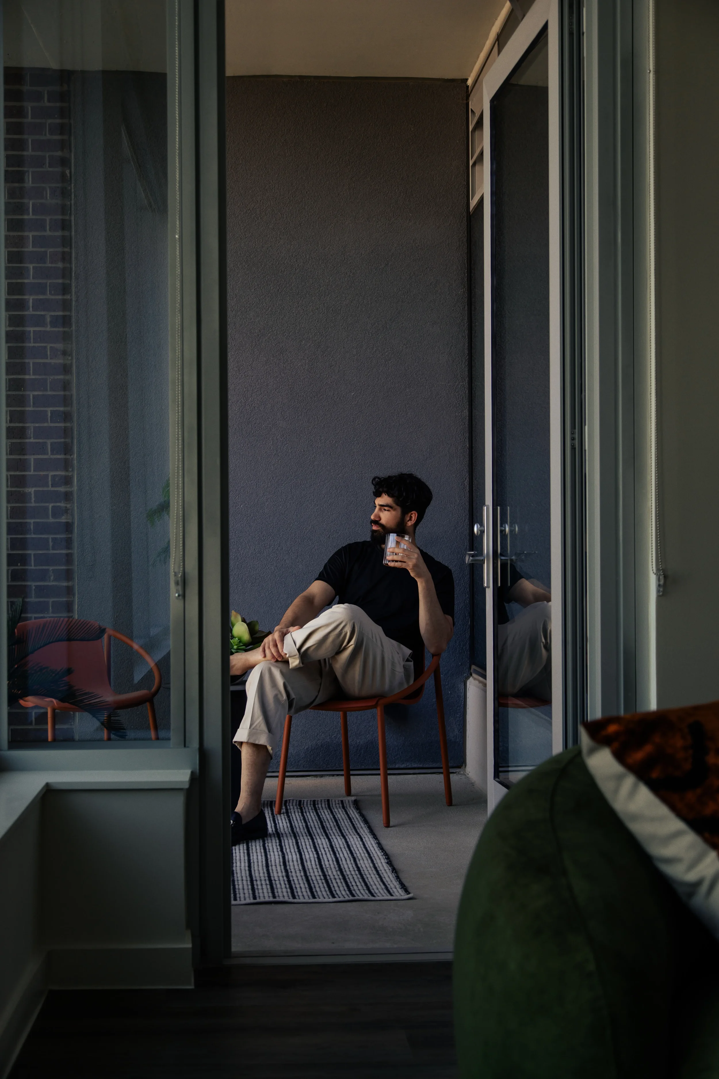 Man sitting on a chair on a balcony or patio holding a drink and looking to the side through an open door frame