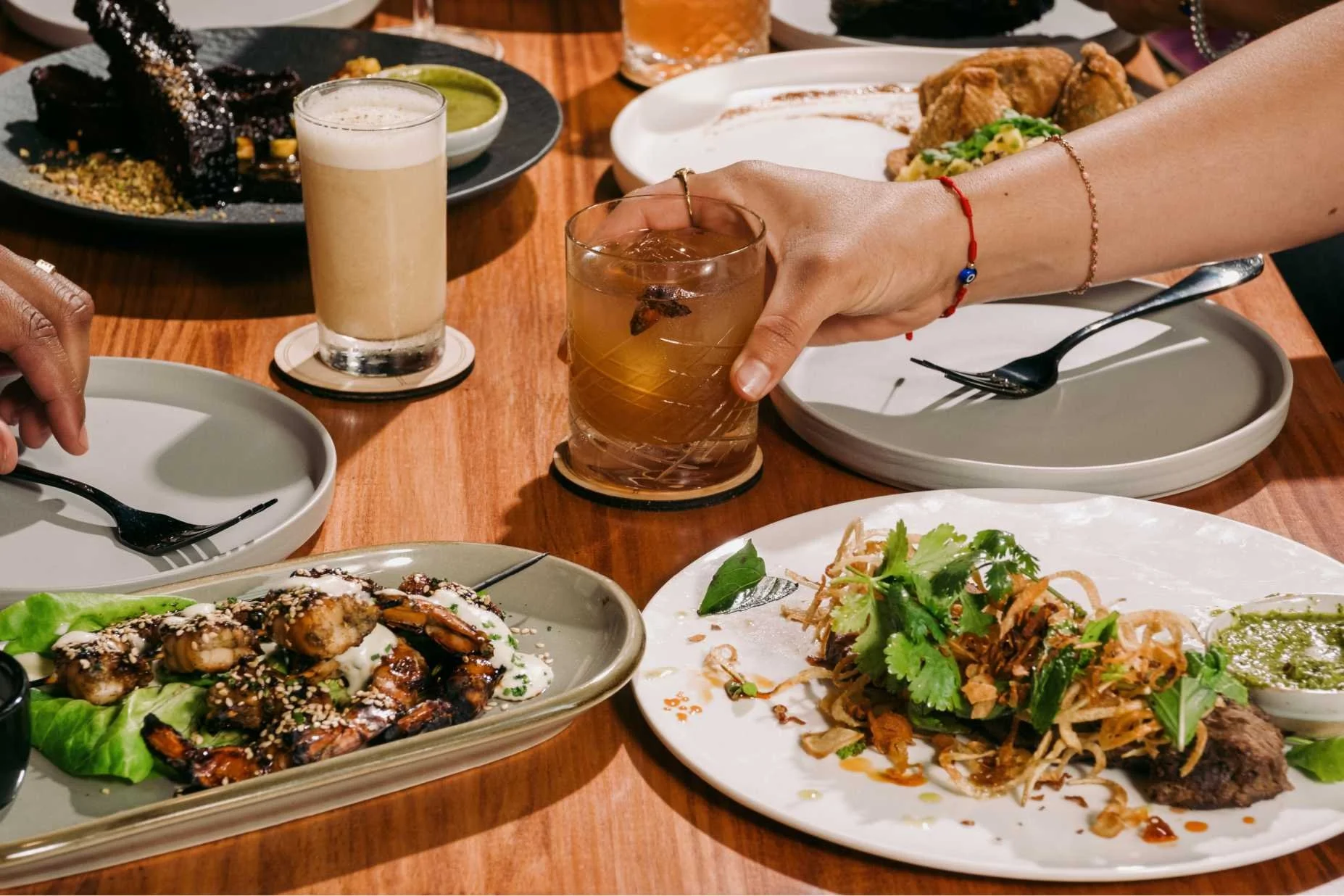Table with various dishes including grilled meat with sesame seeds, pasta with cilantro, and fried chicken with greens, along with two drinks and utensils.