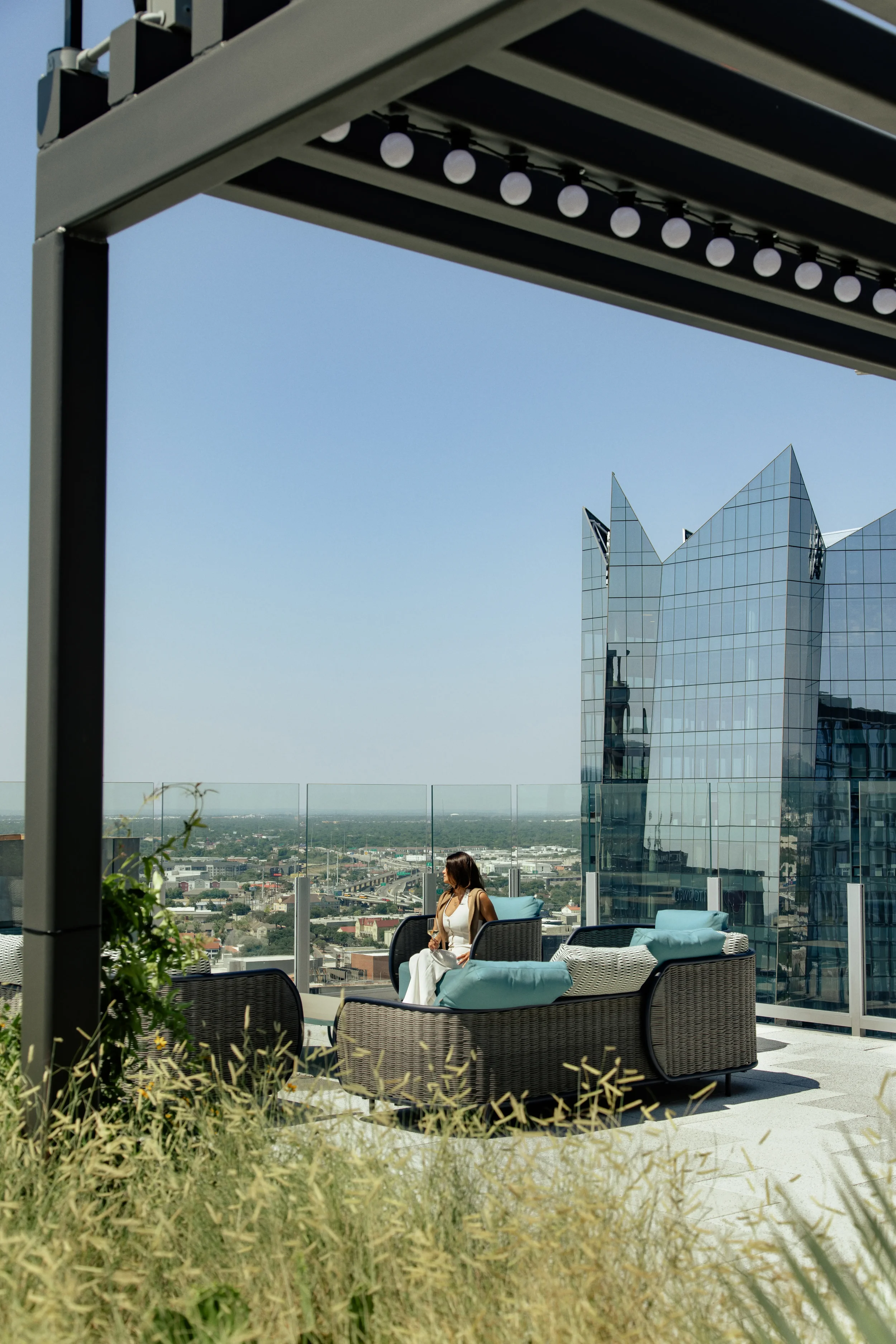 Woman sitting on a rooftop patio with outdoor furniture under a pergola, overlooking a city skyline with a modern glass building in the background