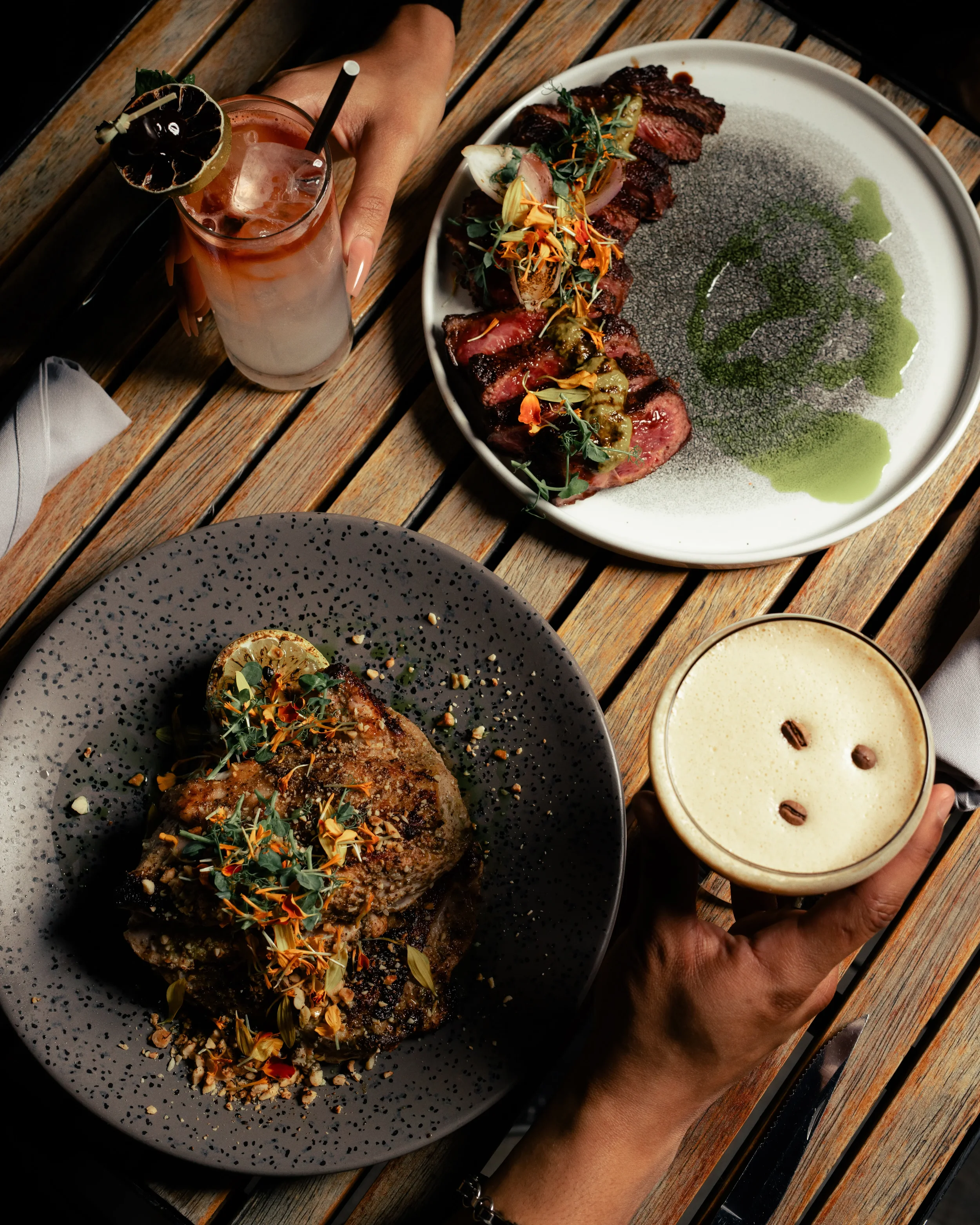 A top-down view of a wooden table set with a cocktail, two plates of steak, and a foamy coffee drink, with two hands holding the drinks.