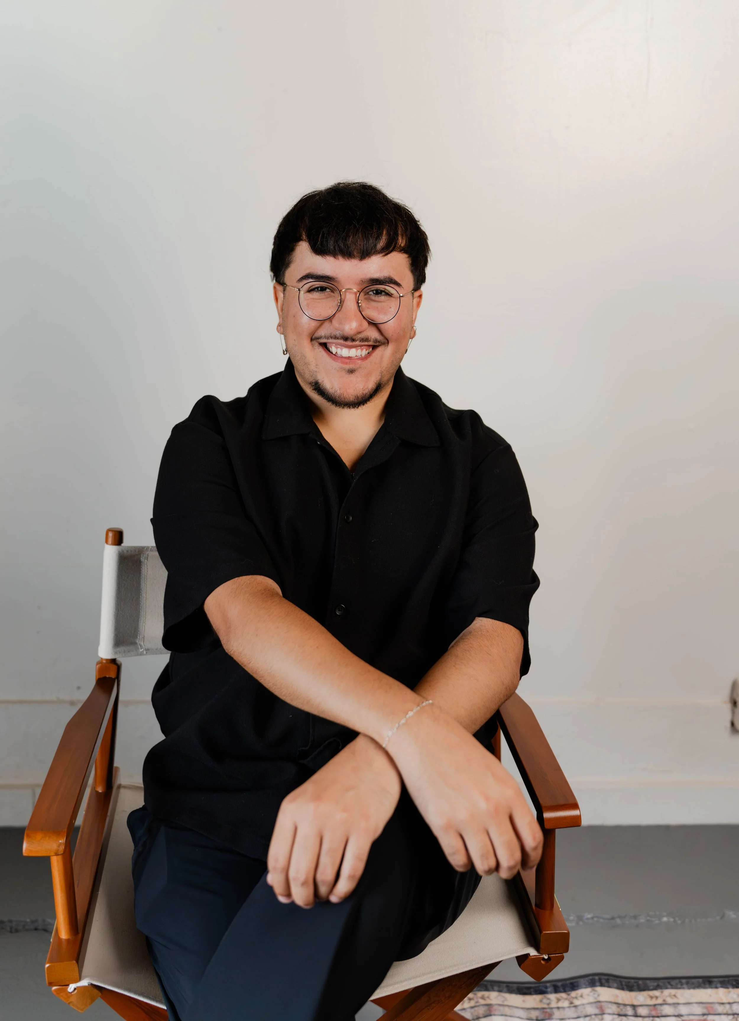 A young man with dark hair and glasses, wearing a black shirt and pants, standing in front of a white brick wall, smiling with one hand resting on his chin.