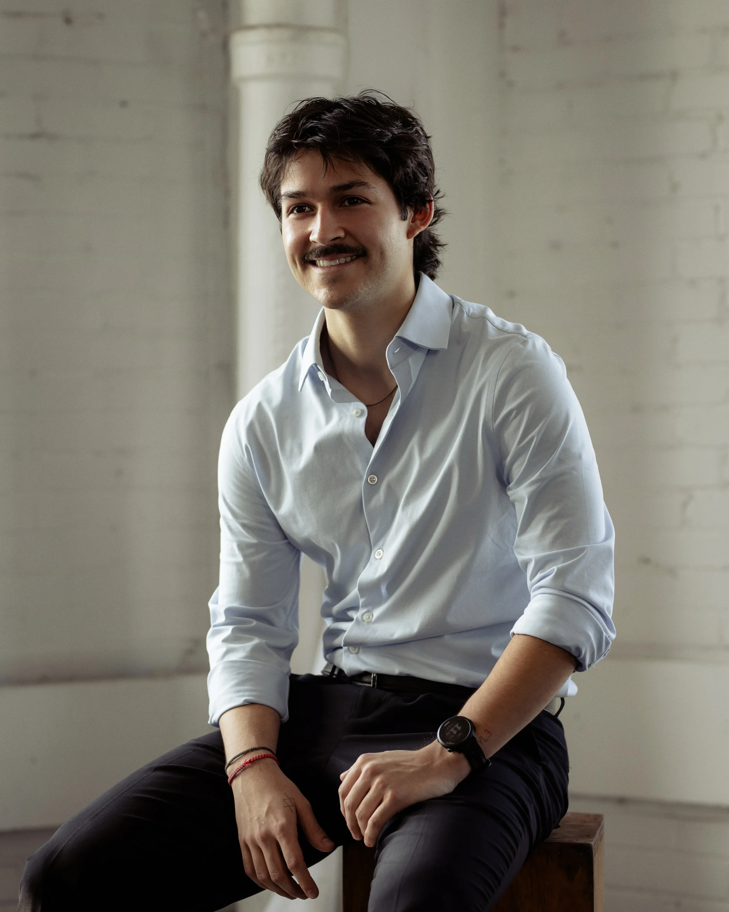 A young man with dark hair, wearing a striped button-up shirt, smiling in a bright indoor setting with a window and potted plant in the background.