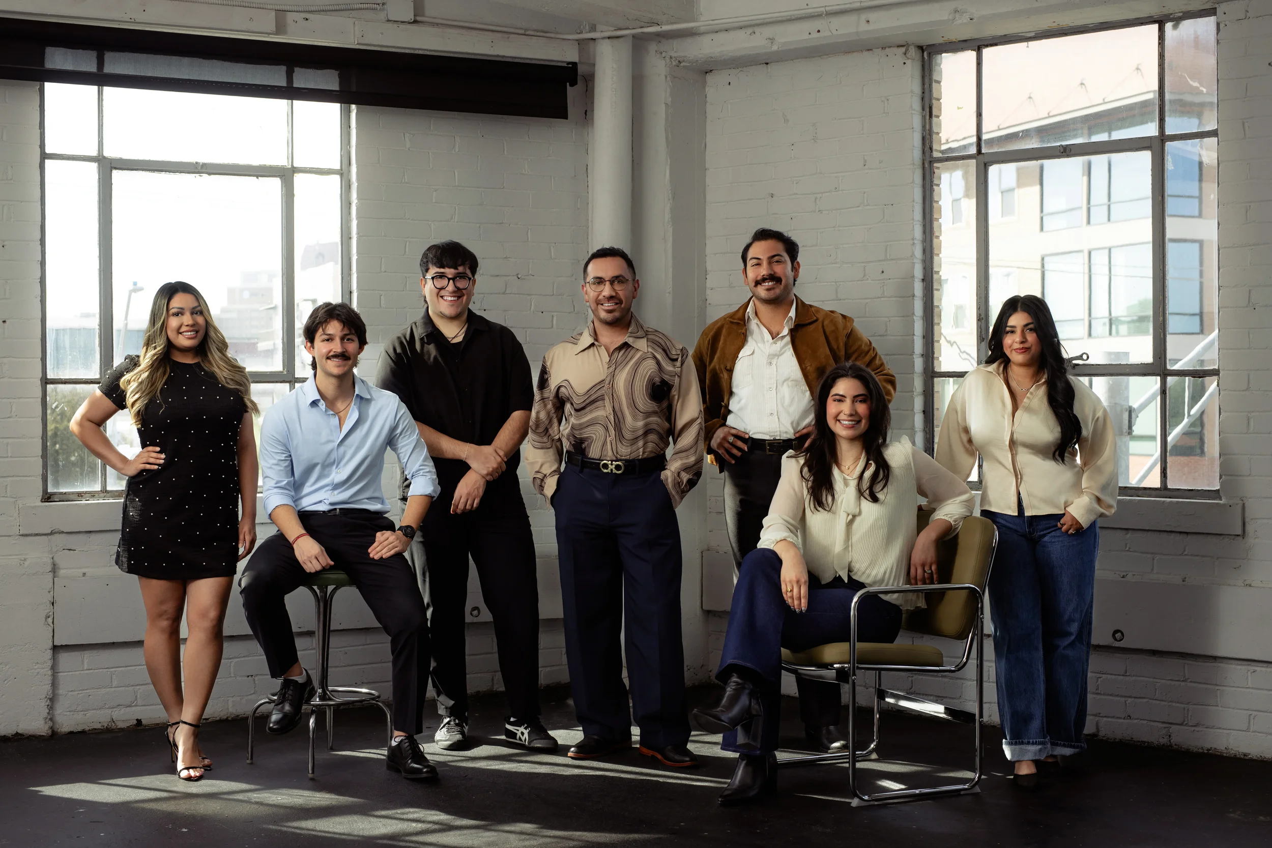 Group of eight diverse young professionals in casual business attire posing in a modern office with large windows and white brick walls.