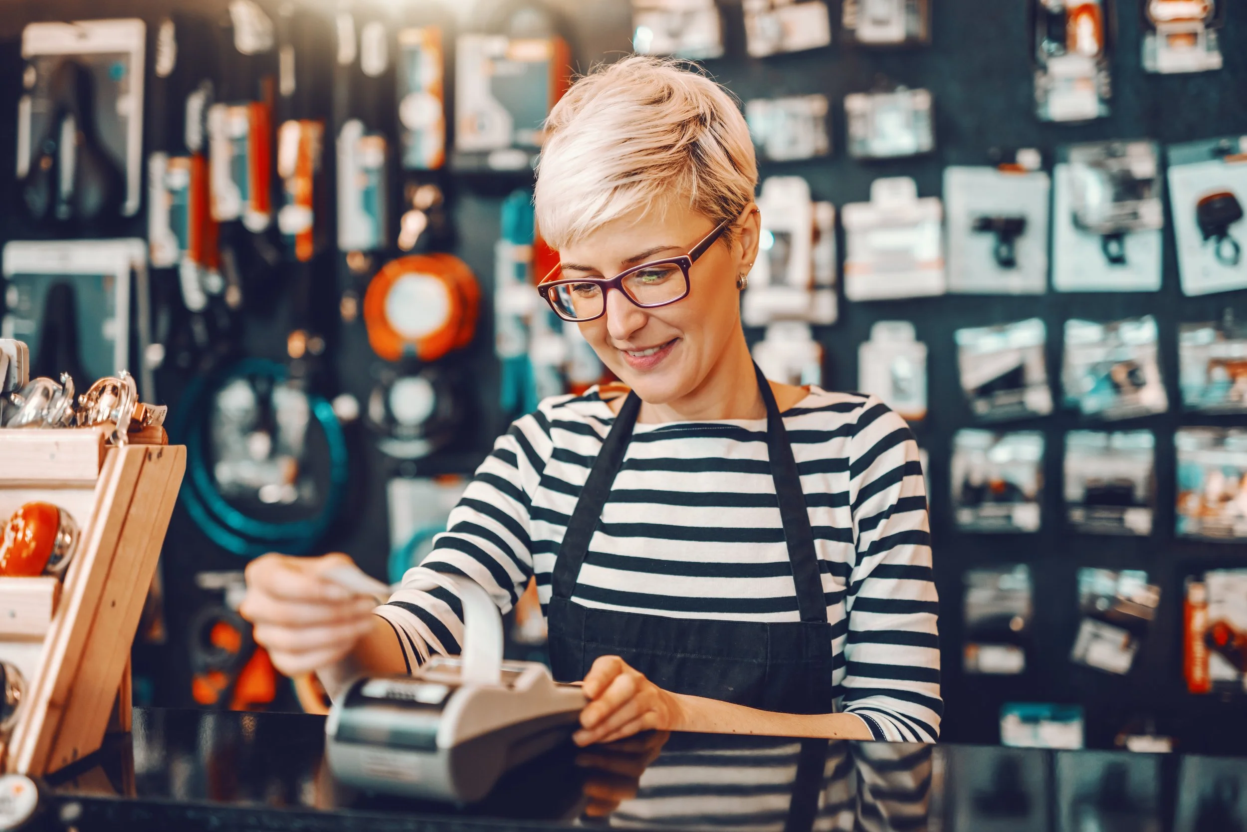 A woman with short blonde hair, glasses, wearing a black and white striped shirt and a black apron, smiling while using a card scanner at a store counter.
