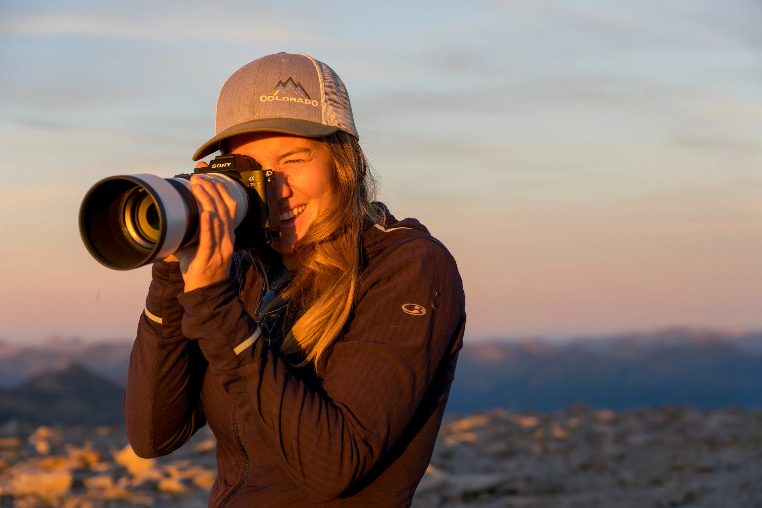 Woman in outdoor gear taking a photograph with a camera during sunset, with a mountain landscape in the background.