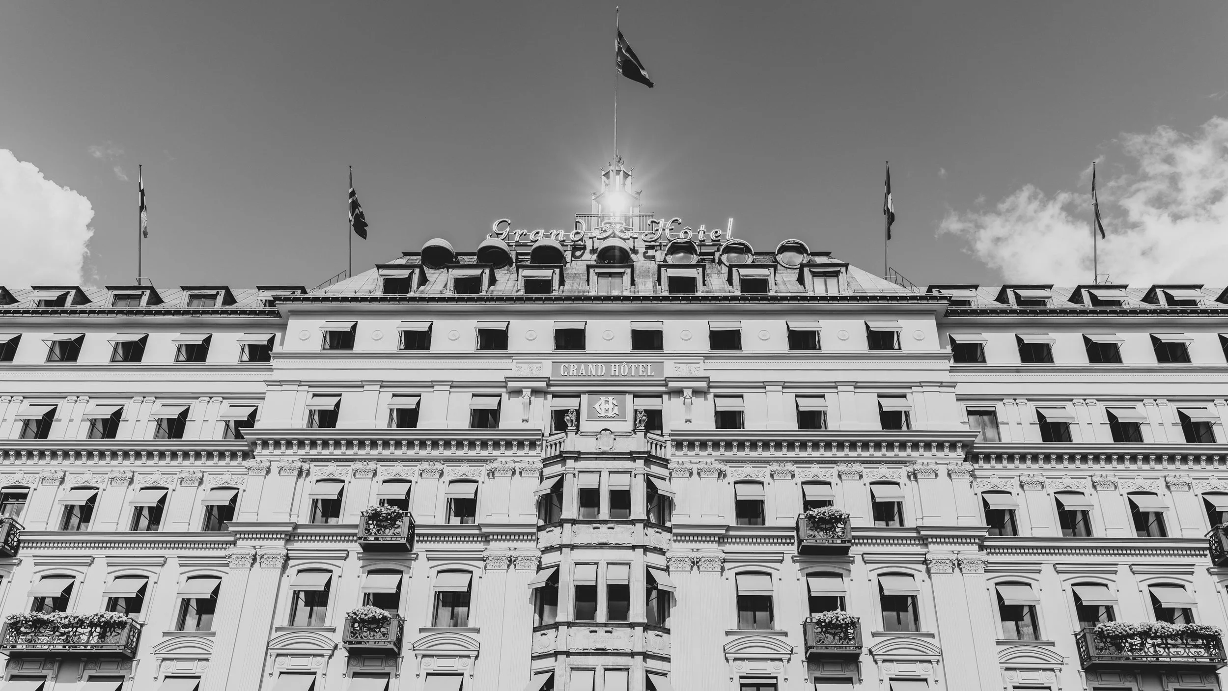 Front view of a large, historic hotel building called Grand Hotel, with multiple windows, balconies, and flags on top, in black and white photography.