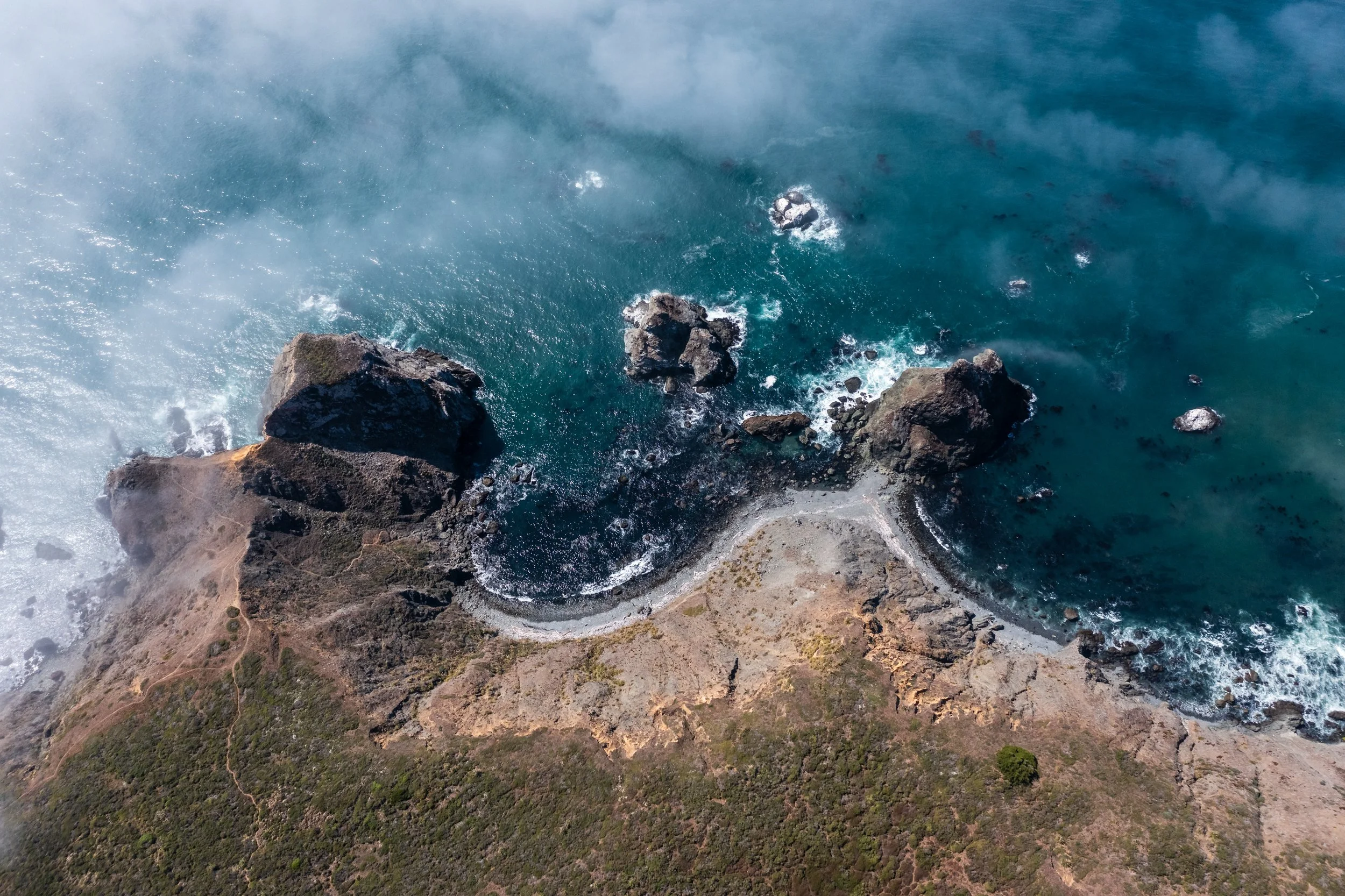 Aerial view of rocky coastline with waves crashing against rocks and a curved dirt path along the land.