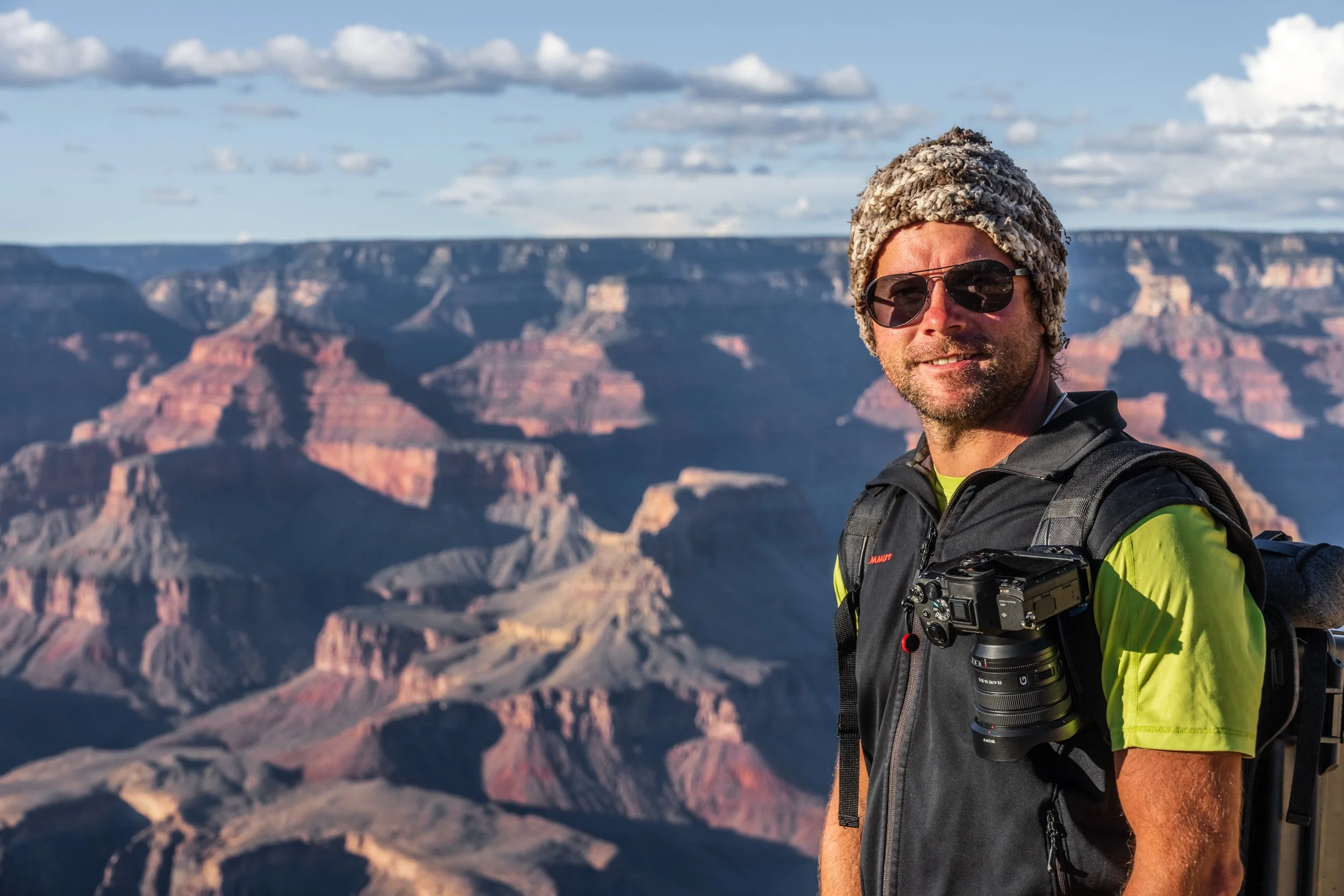 A man with sunglasses, a knit hat, and a bright yellow shirt stands in front of the Grand Canyon, with a camera hanging from his neck and a backpack on his shoulder.