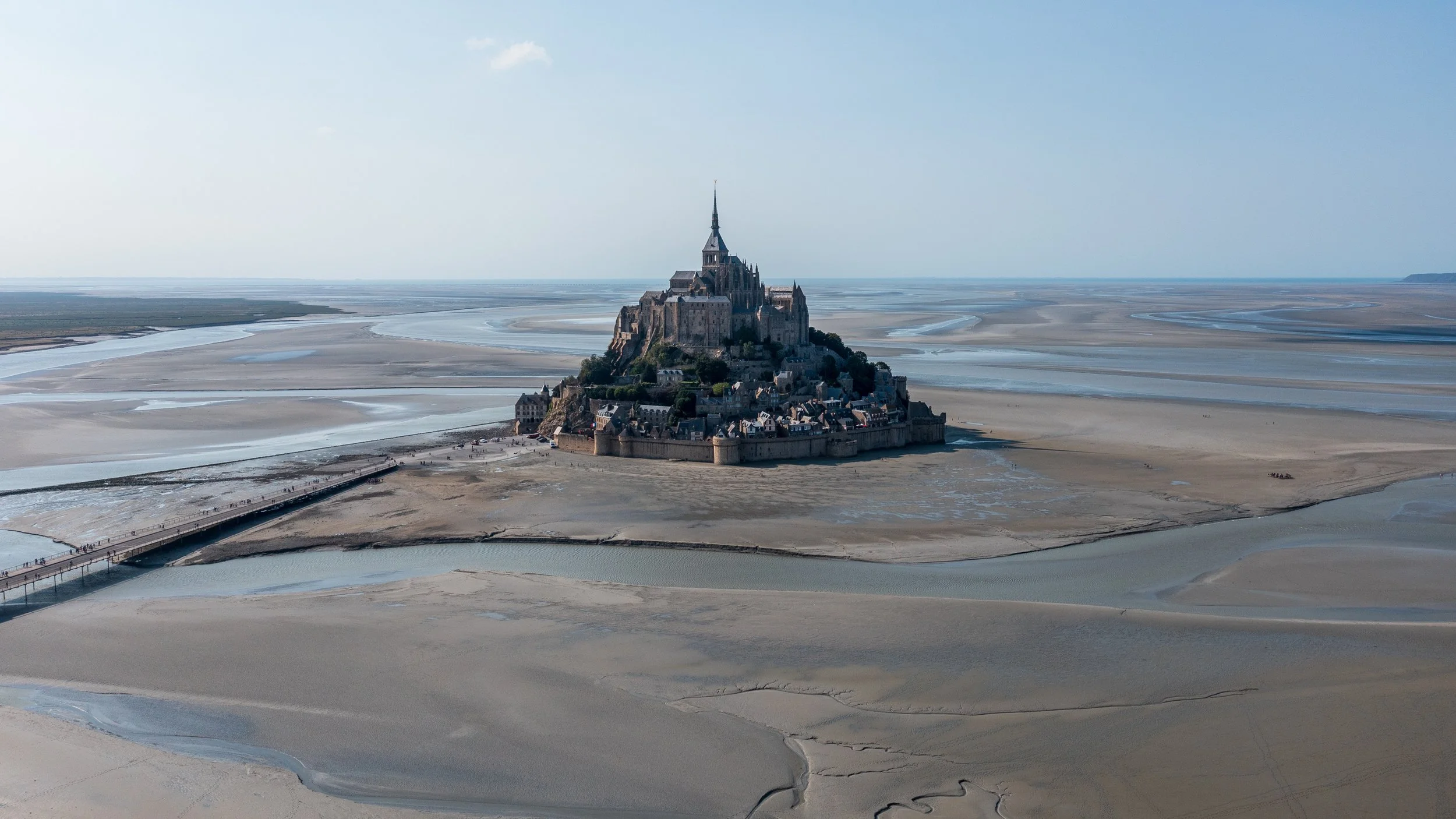 A large island with a historic castle and village, surrounded by water and sand in a coastal area, with a bridge connecting to the mainland.