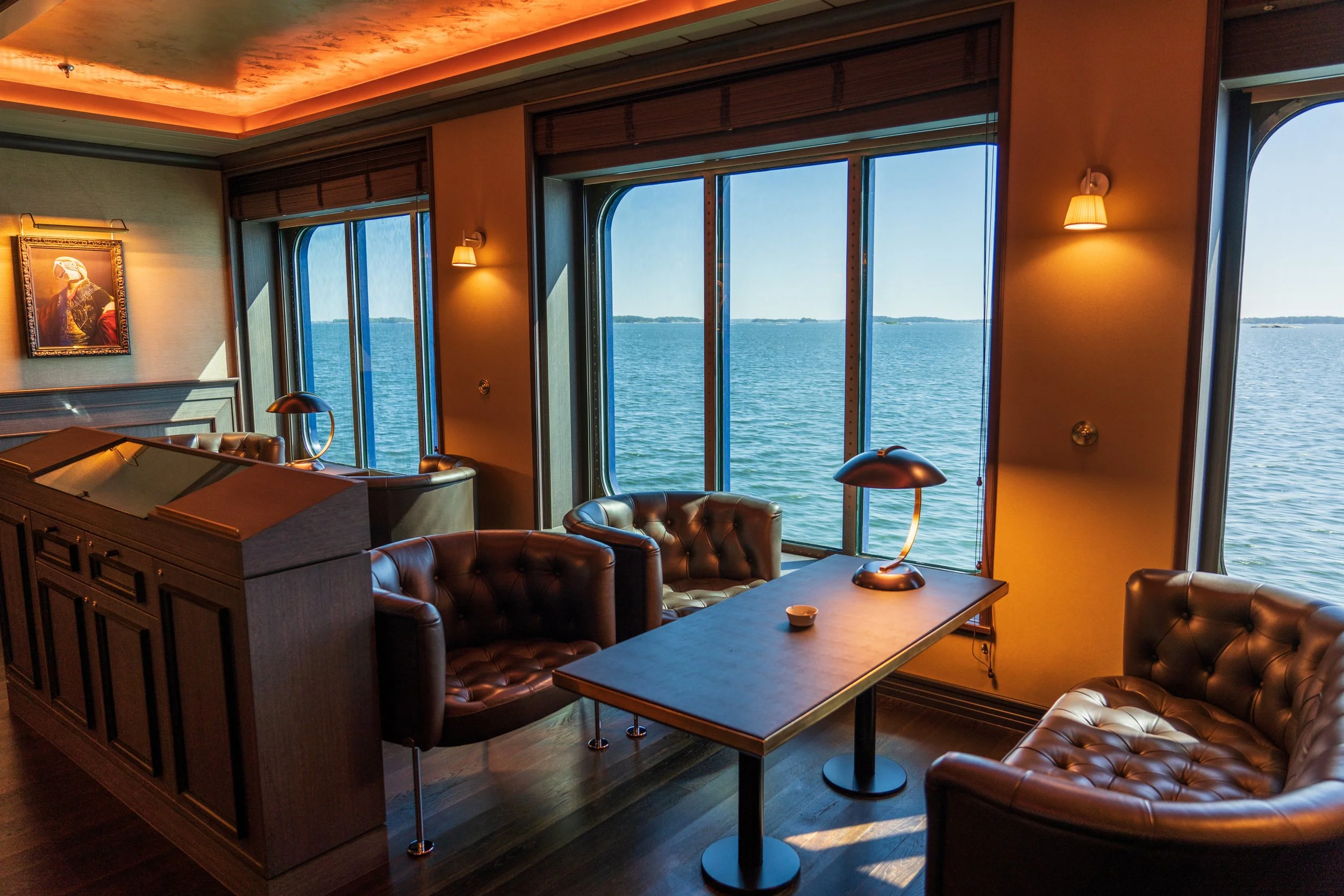 Interior of a room with large windows overlooking the water, leather chairs, a wooden table with a small lamp, and warm lighting.