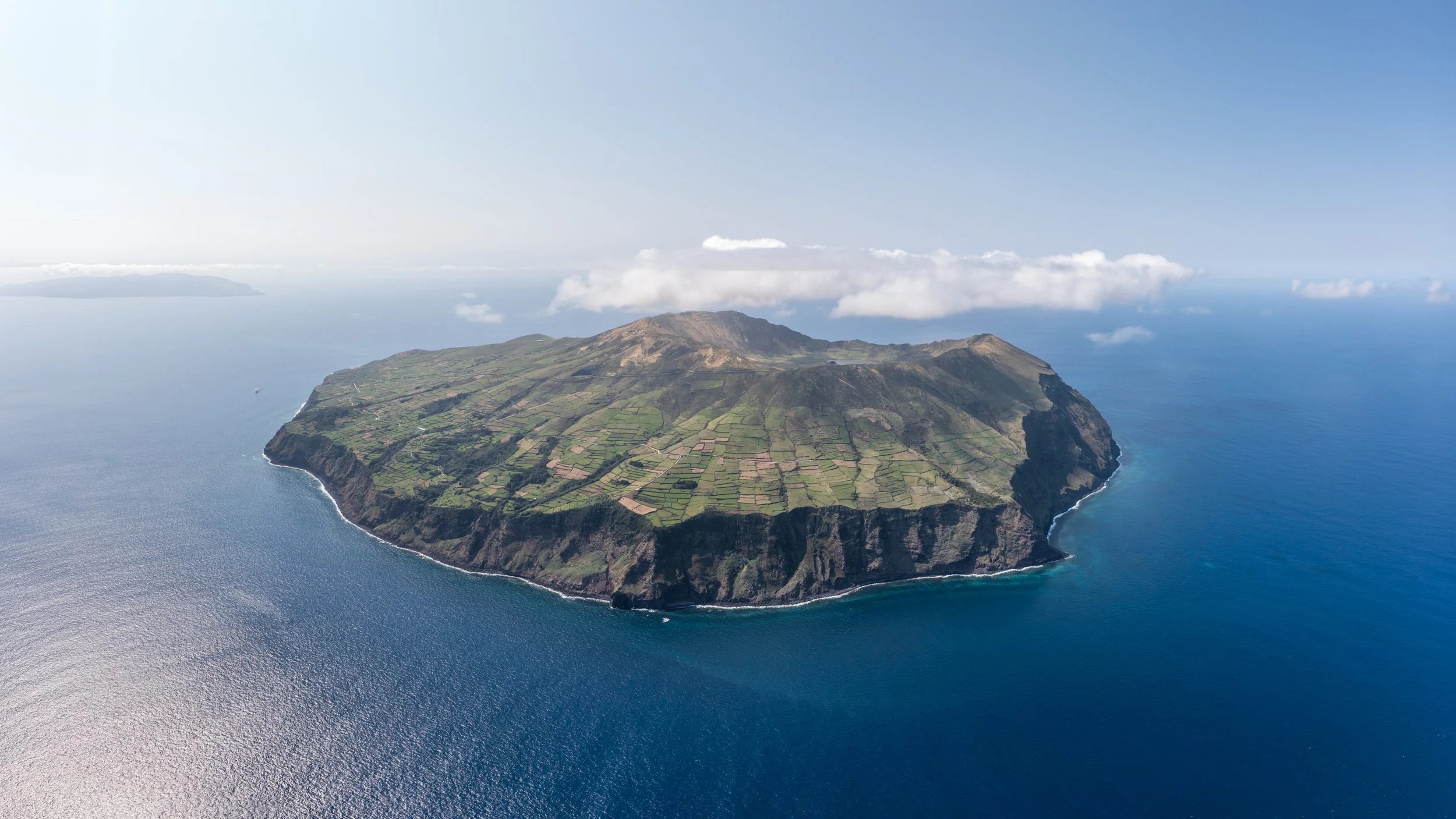 Aerial view of a volcanic island with lush green terraced fields, surrounded by blue ocean, under a partly cloudy sky.