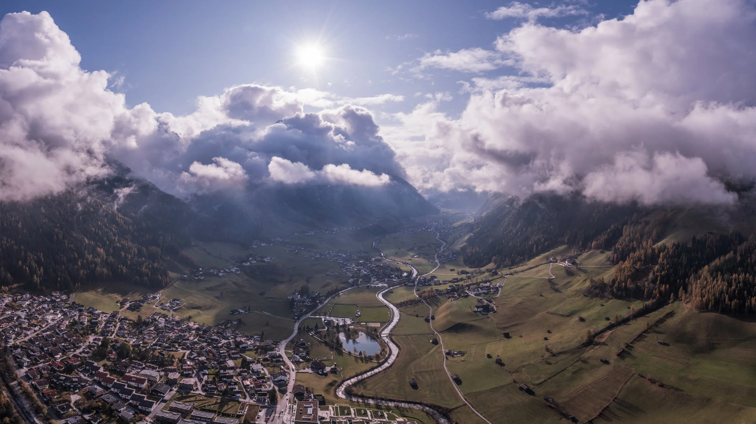 Aerial view of a small town in a valley surrounded by mountains with clouds above and the sun shining brightly.