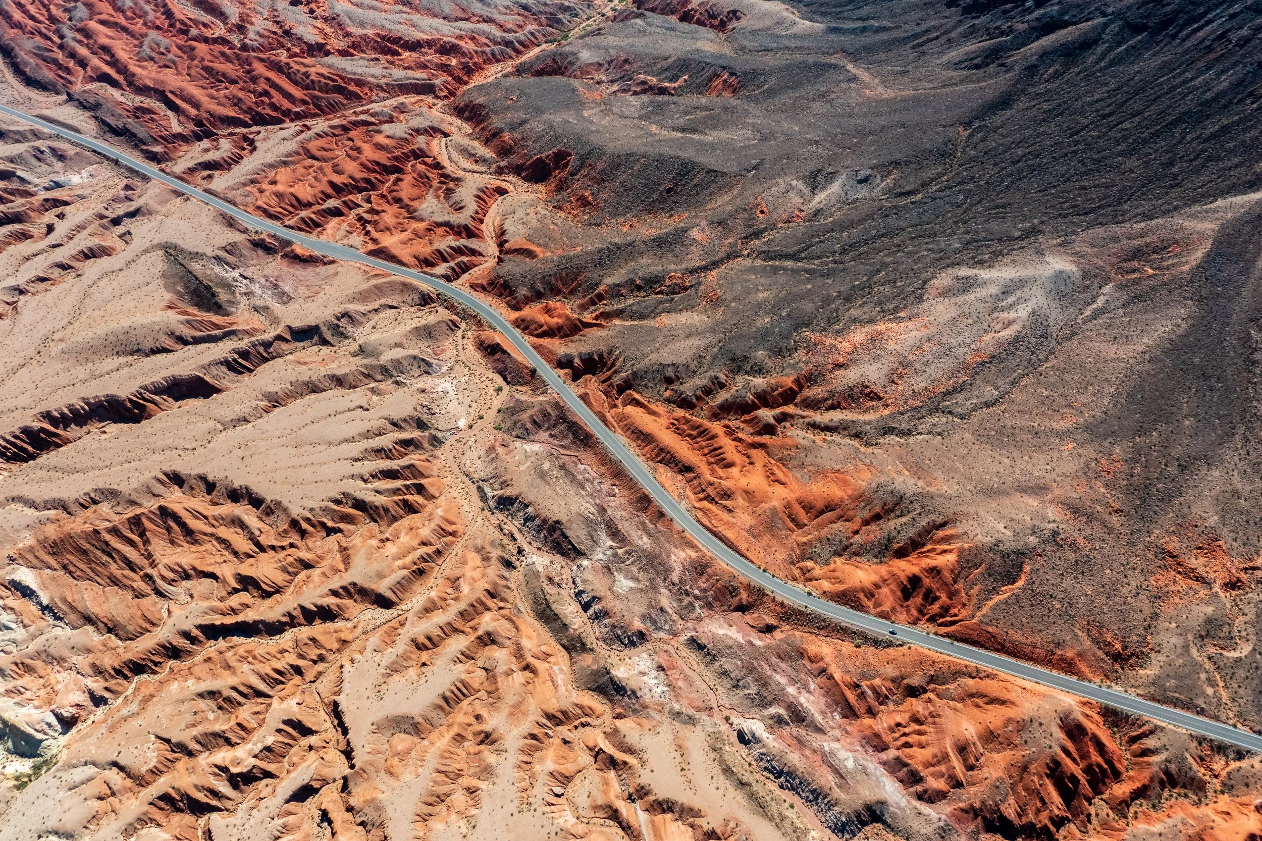 Aerial view of a winding road through a desert landscape with red and brown rock formations and sparse vegetation.