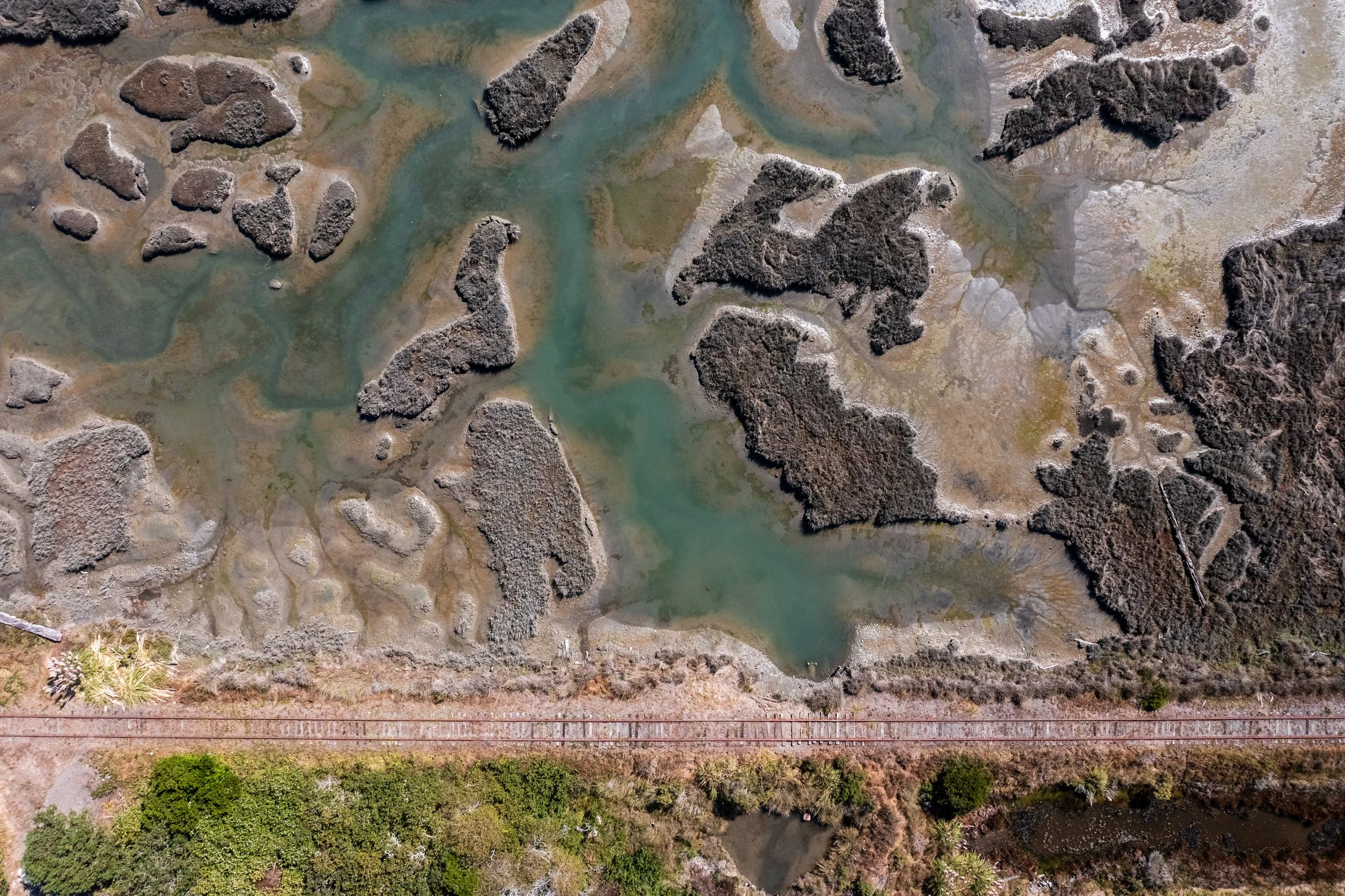 An aerial view of a marshland with winding rivers, clusters of grass and shrubs, and a nearby railroad track at the bottom.