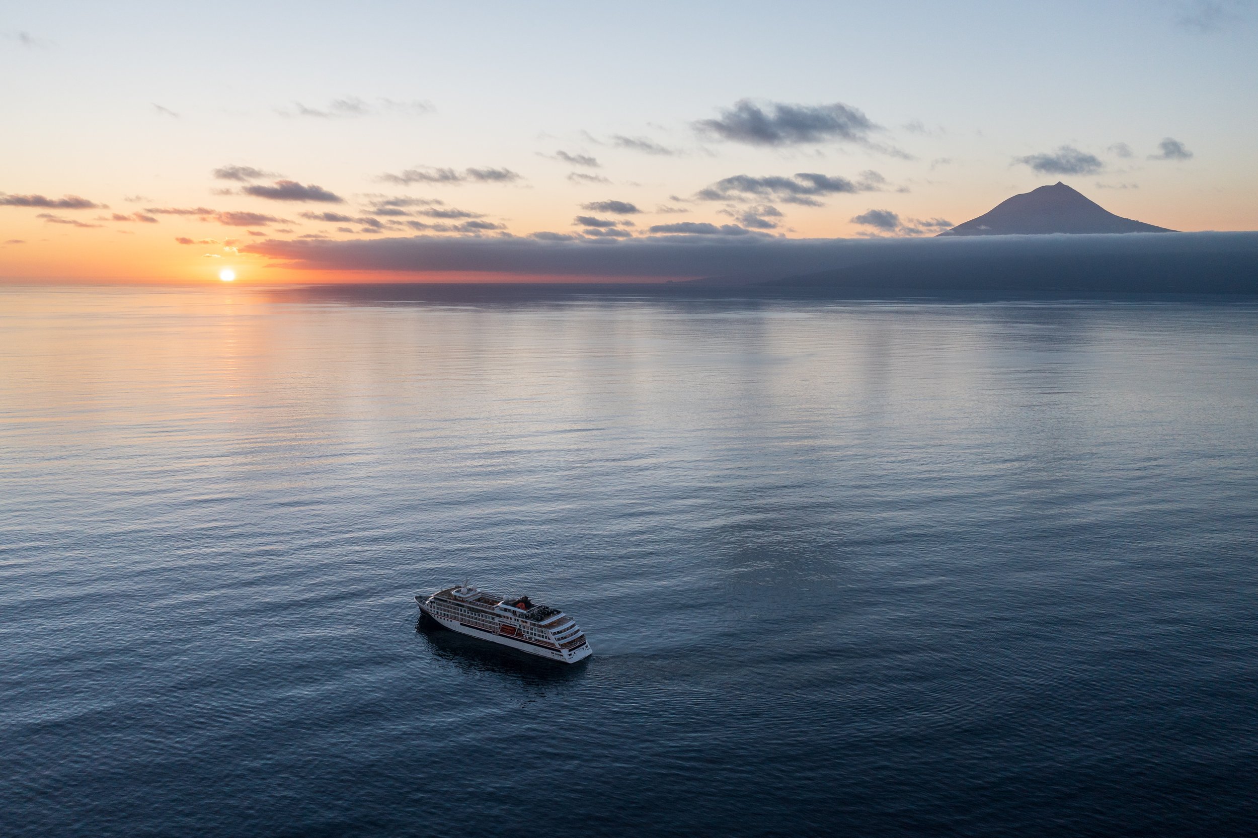 A cruise ship in calm ocean waters during sunset, with a volcano in the distance and a few scattered clouds in the sky.