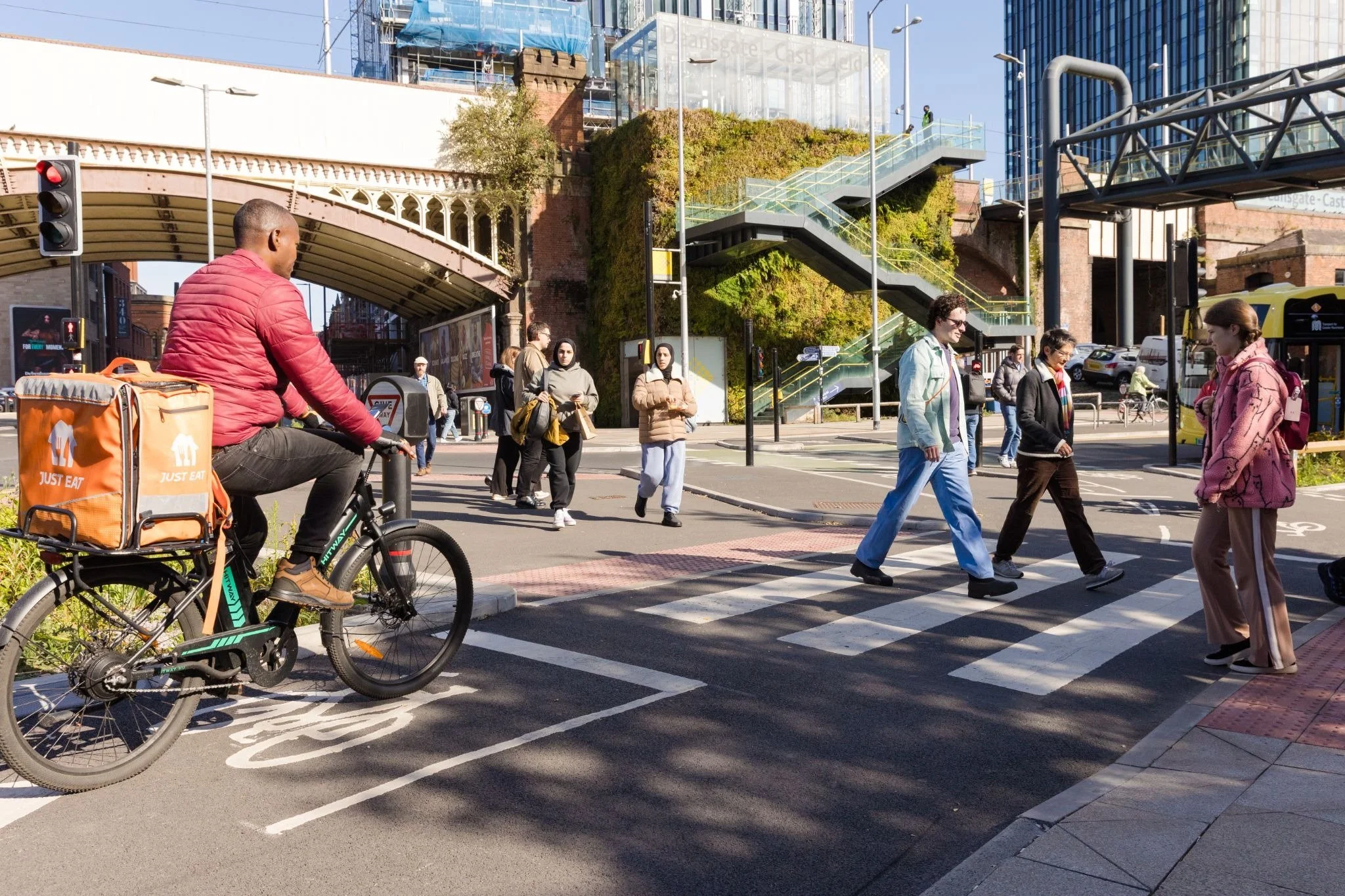 Alt text: A cyclist waits at a cycle lane as multiple pedestrians cross. The image is taken on a sunny day on Deansgate in Manchester