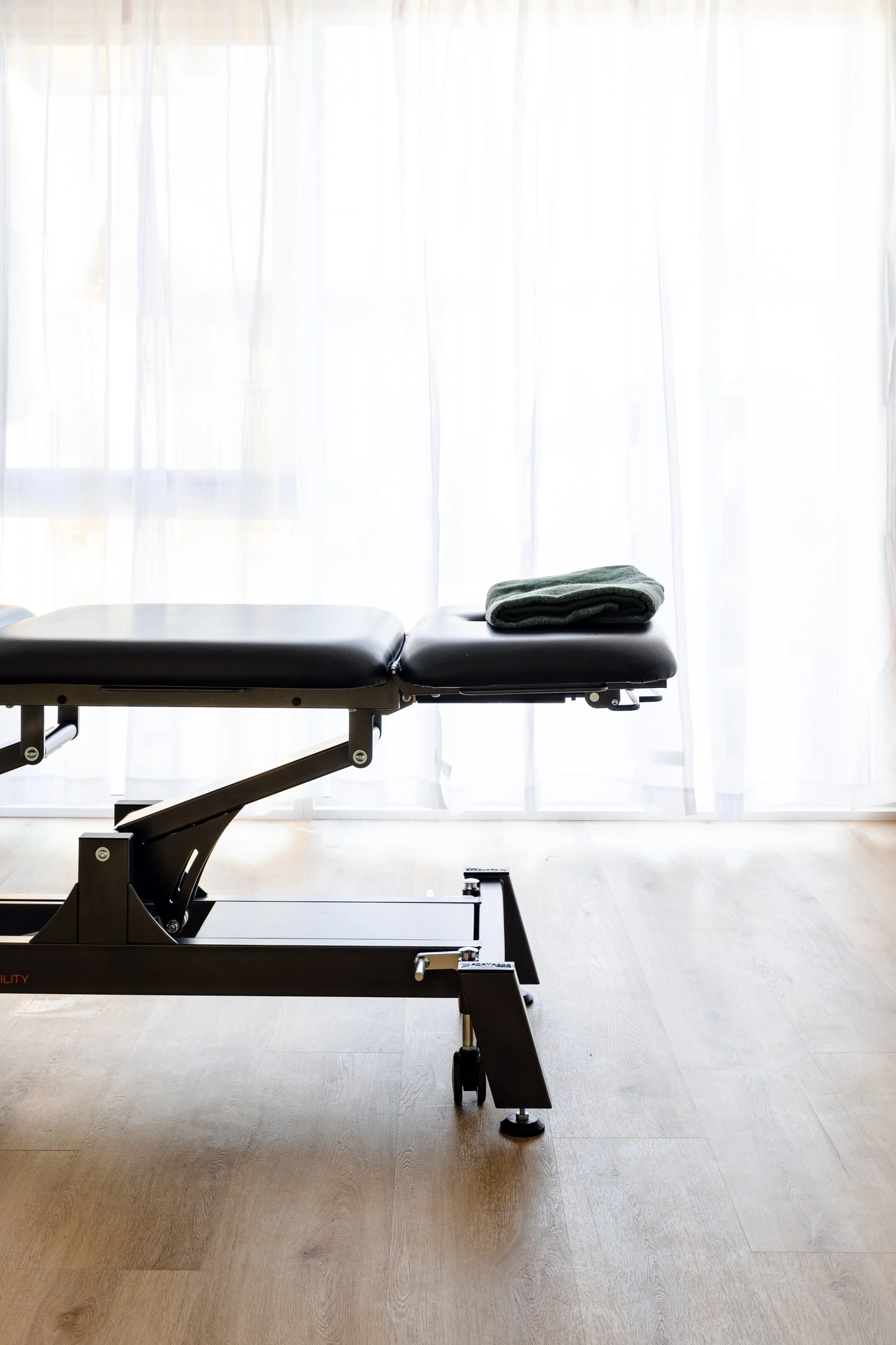 Empty massage table with folded towel on top, in front of a sheer curtain background.