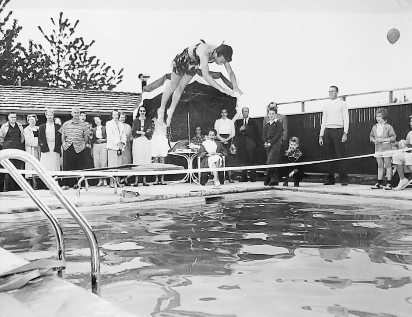 Historical black and white of person jumping off diving board into pool with onlookers on the pool deck