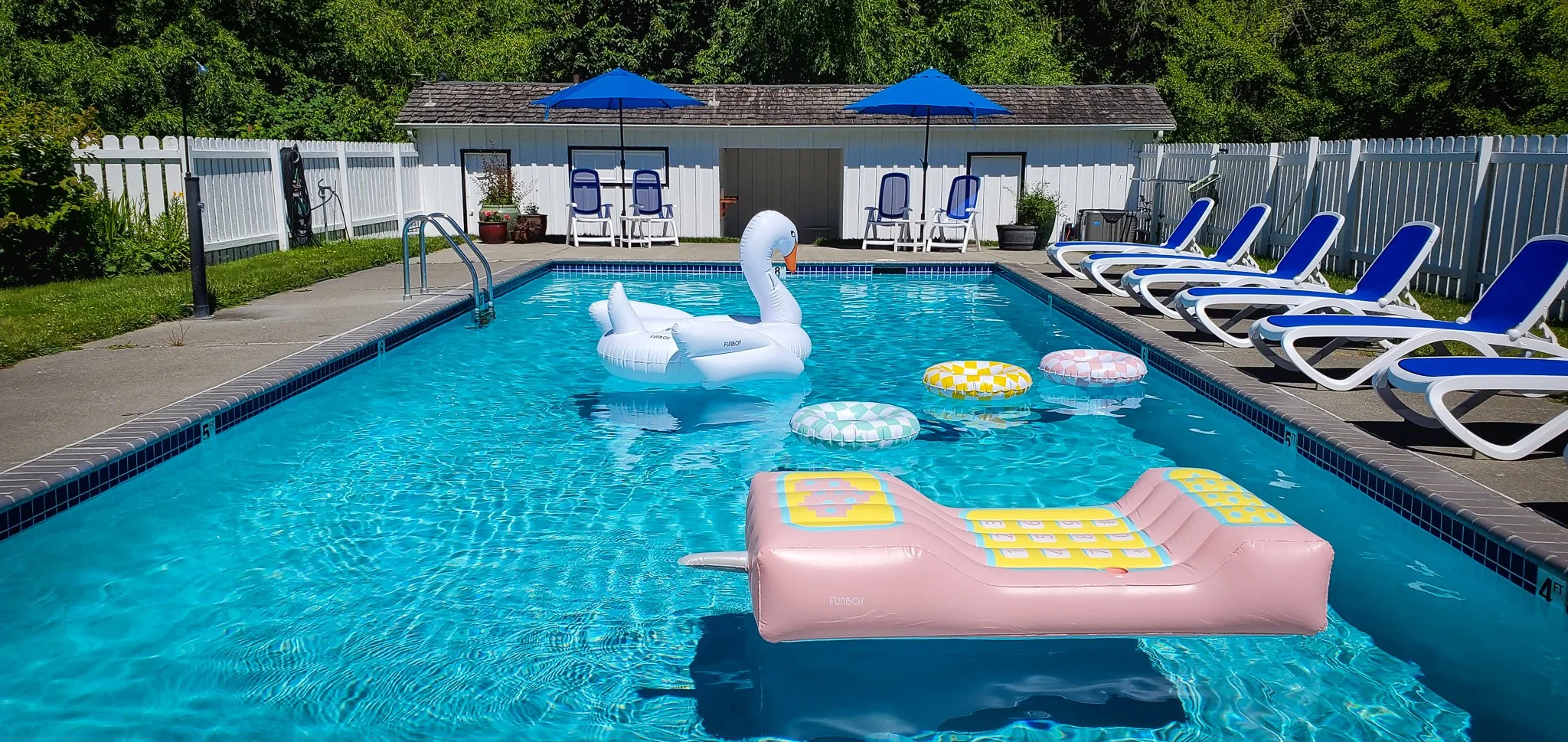 Beautiful blue pool with white swan, pink phone and other floaties with white pool house with black trim and blue chairs and umbrellas on the pool deck.