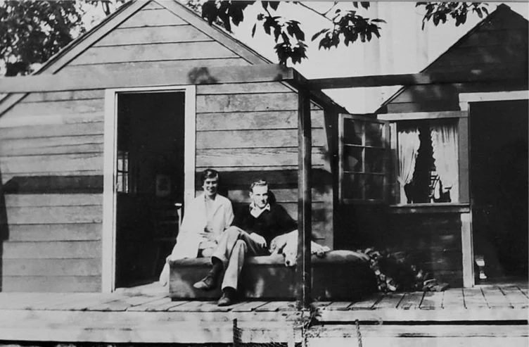Historical black and white of two cabins nex to eachother with couple and their pup sitting on bench outside