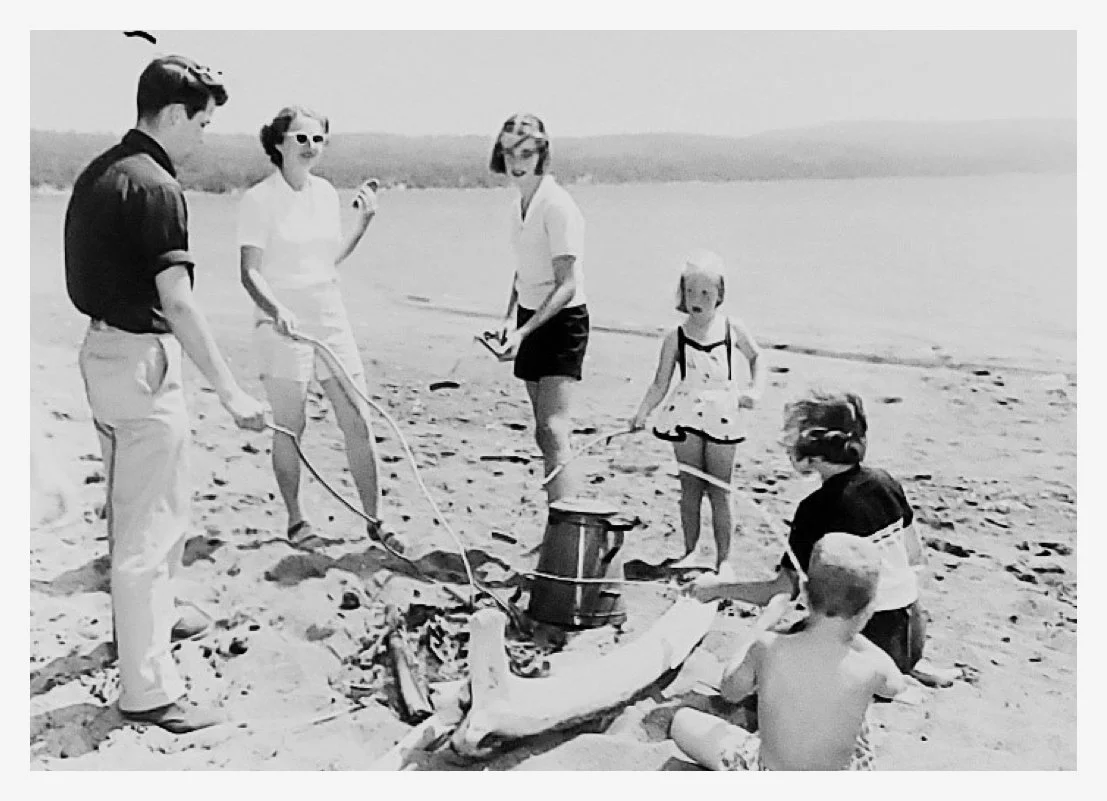 Historcial black and white of young family of six at the beach in the summer roasting hotdogs on sticks over a campfire