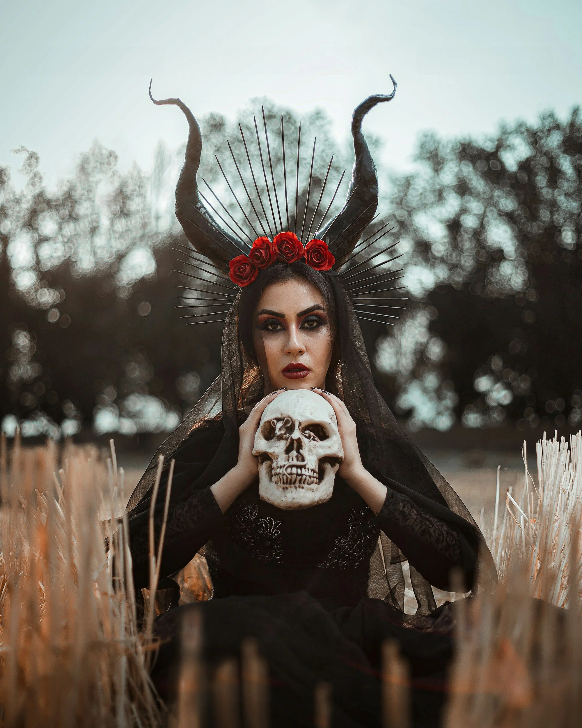 Woman in black attire with horns and roses, holding a skull in a field.
