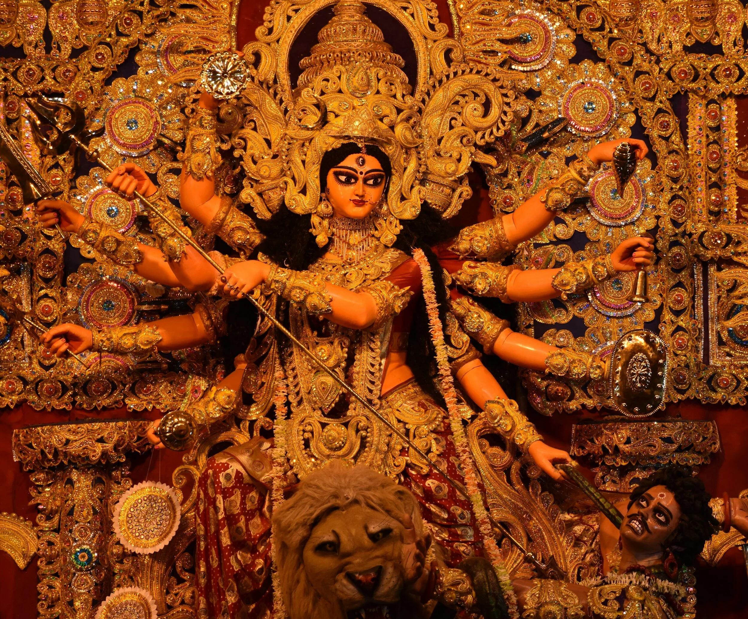 Statue of Hindu goddess Durga with multiple arms holding weapons, adorned in intricate gold decorations, with a lion at her feet and a defeated demon."
