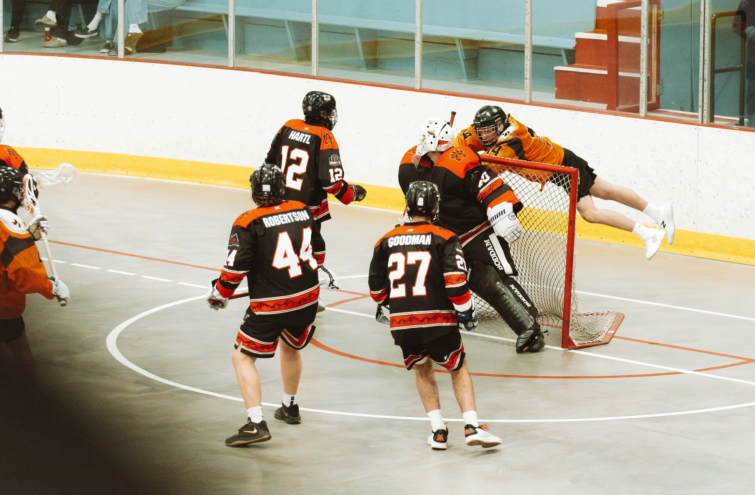 Kodiaks box lacrosse players Hartl, Robertson, and Goodman watch as their goalie defends the net while an opposing player in orange leaps through the air during a game.