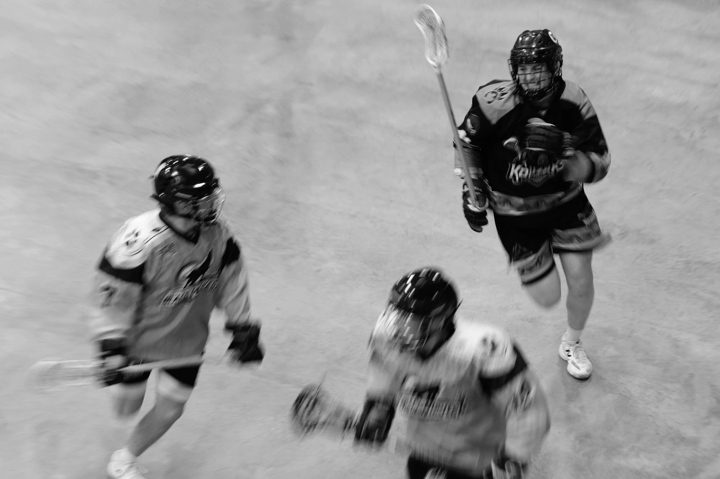Black and white action photo of a Kodiaks box lacrosse player running with his stick as two opposing players move across the arena floor.