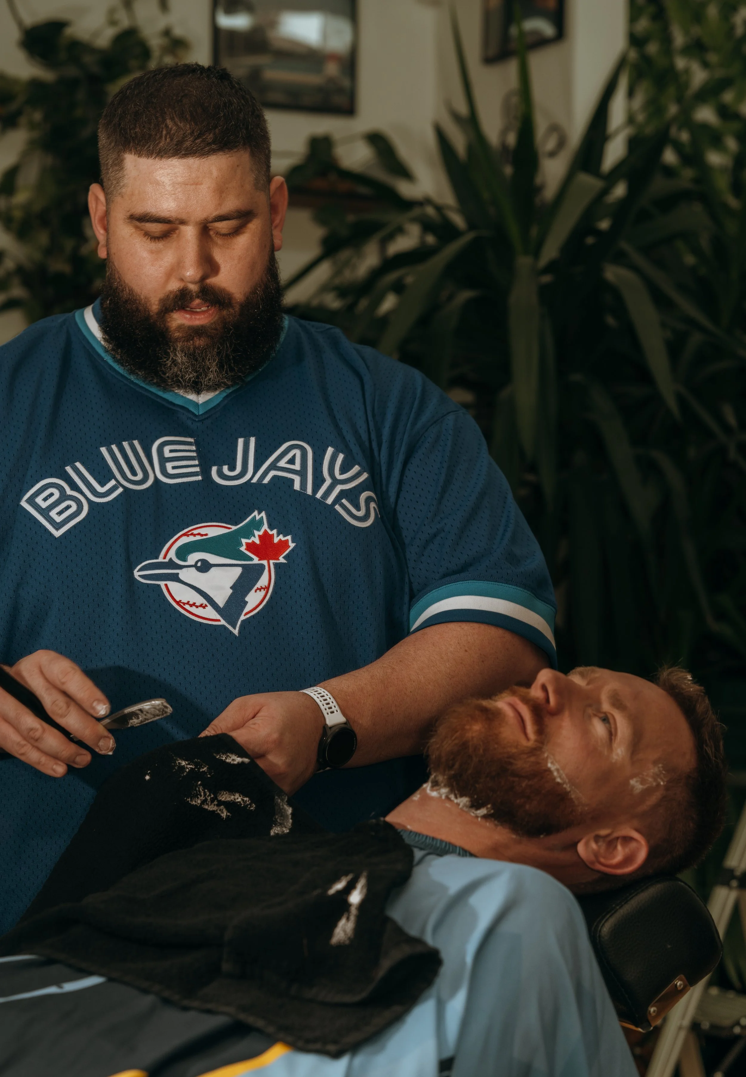 A man getting a straight razor shave at a barbershop. The barber, wearing a Toronto Blue Jays jersey, is holding the man's chin with one hand and a razor with the other. The man is reclining with a towel on his chest, and appears relaxed.