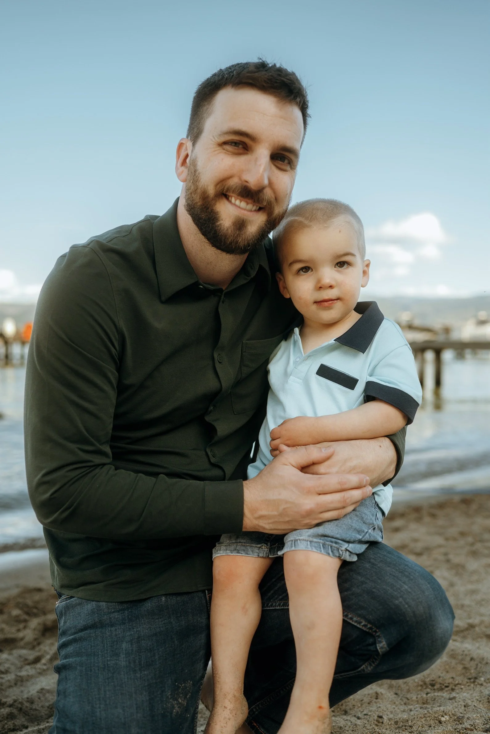 A man with a beard smiling at the camera while holding a young boy with short hair at the beach. The man is wearing a dark green shirt, and the boy is wearing a light blue polo shirt. The background features a pier extending into the water, with a cl