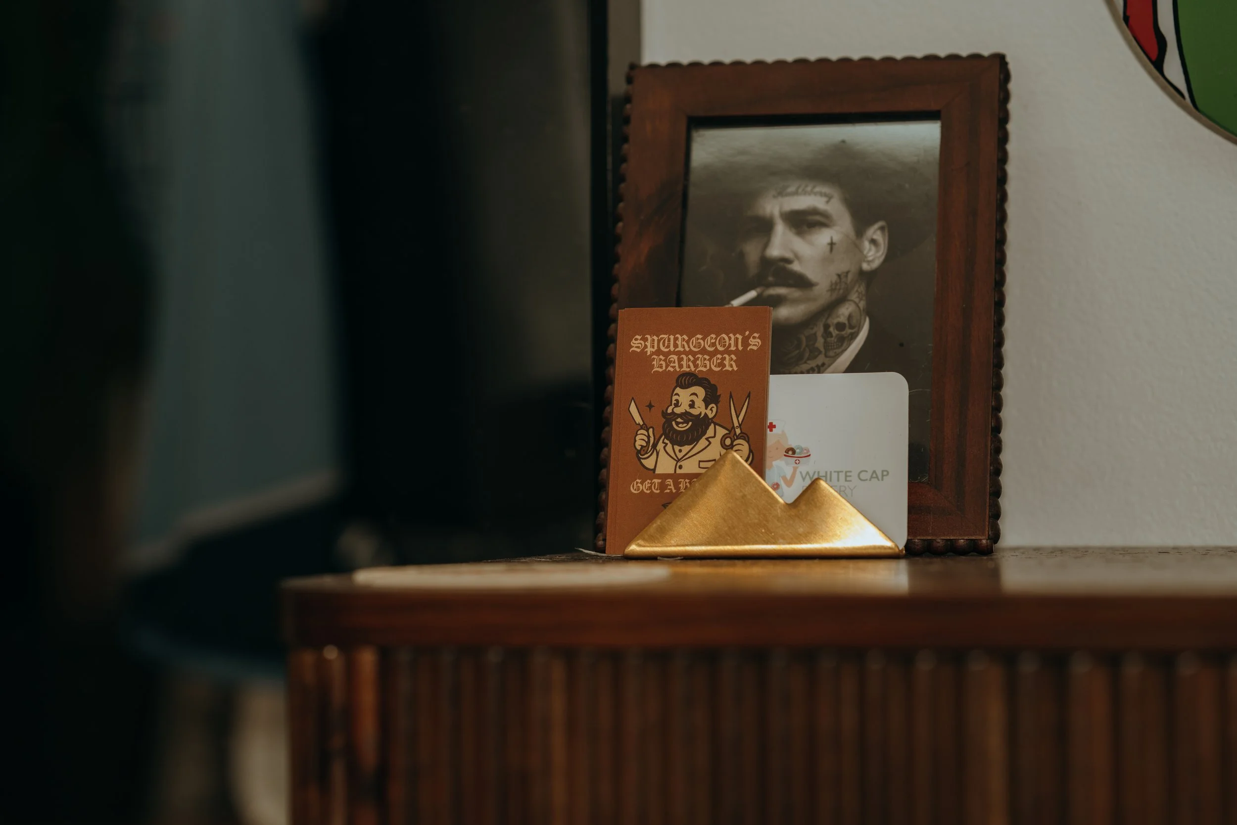 A framed black-and-white photograph of a man with tattoos, a mustache, and a cigarette in his mouth, sitting on a wooden surface. In front of the photo, there are small items including a card with a cartoon barber holding scissors and a comb, a white