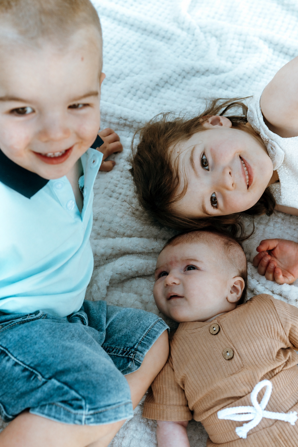 Three children, a boy, a girl, and a baby, lying on a white blanket, smiling and looking at the camera.