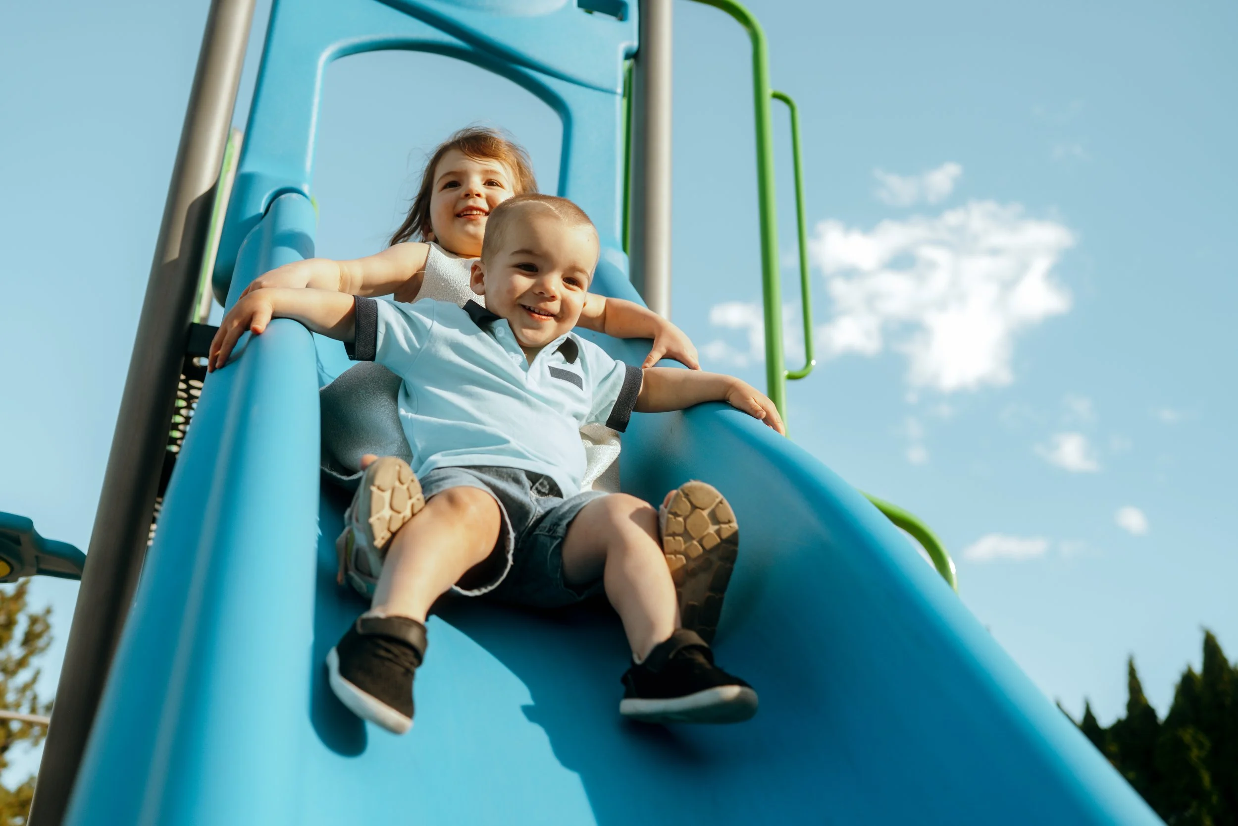 Two young children, a girl with brown hair and a boy with a buzz cut, are riding down a blue playground slide, smiling and enjoying a sunny day outdoors.