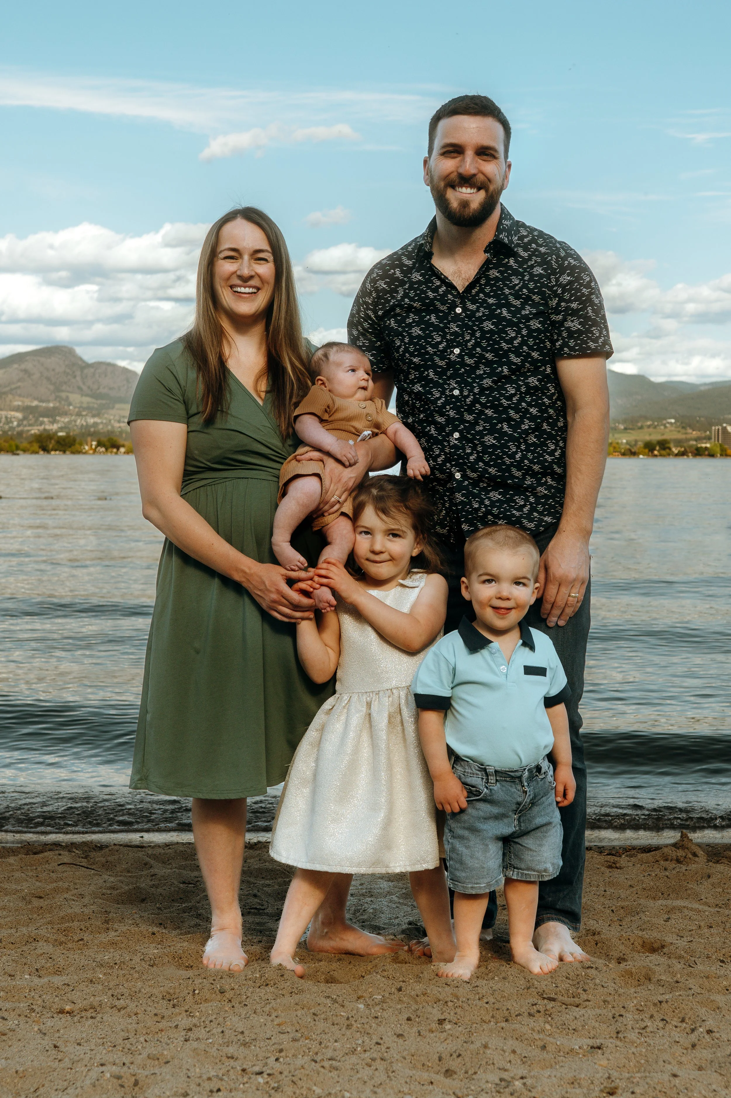 Family of five standing on a sandy beach by a lake with mountains in the background, smiling at the camera.