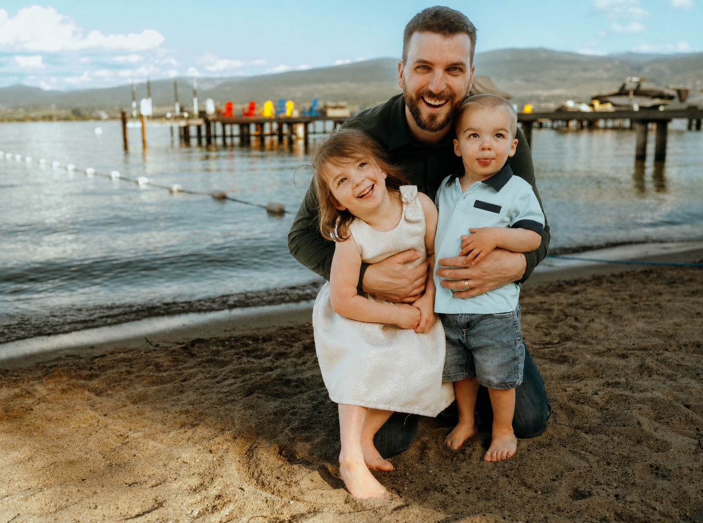 A man with two young children smiling at the camera on a sandy beach with a lake, a pier, and mountains in the background.
