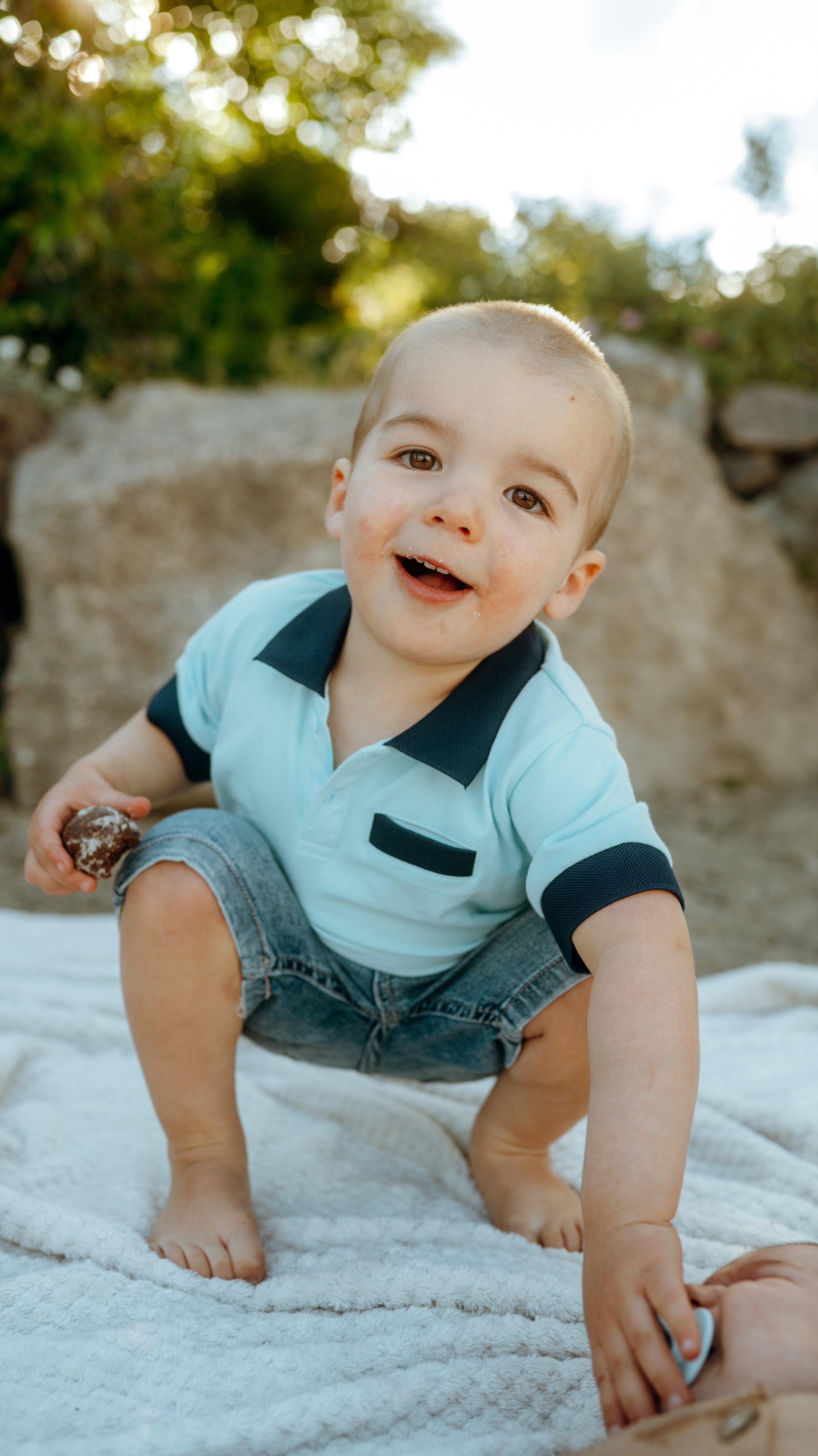 Young boy with short hair, wearing a light blue polo shirt with black accents, crouching on a white blanket outdoors, holding a chocolate snack in one hand and reaching for a shoelace with the other, with a background of trees and sunlight.
