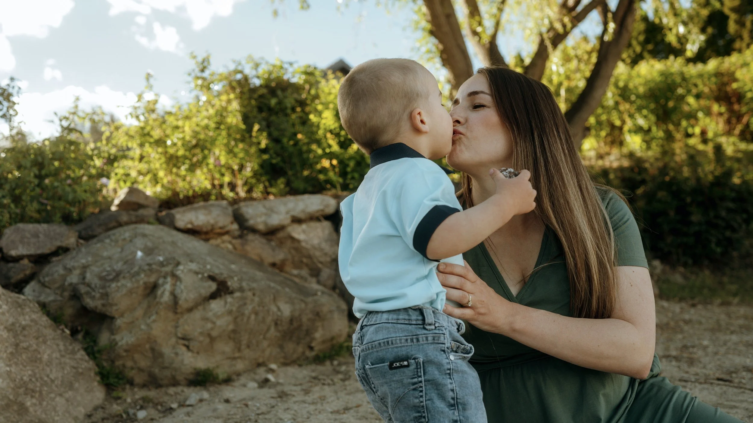 A woman and a young child sharing a kiss outdoors with trees and rocks in the background.