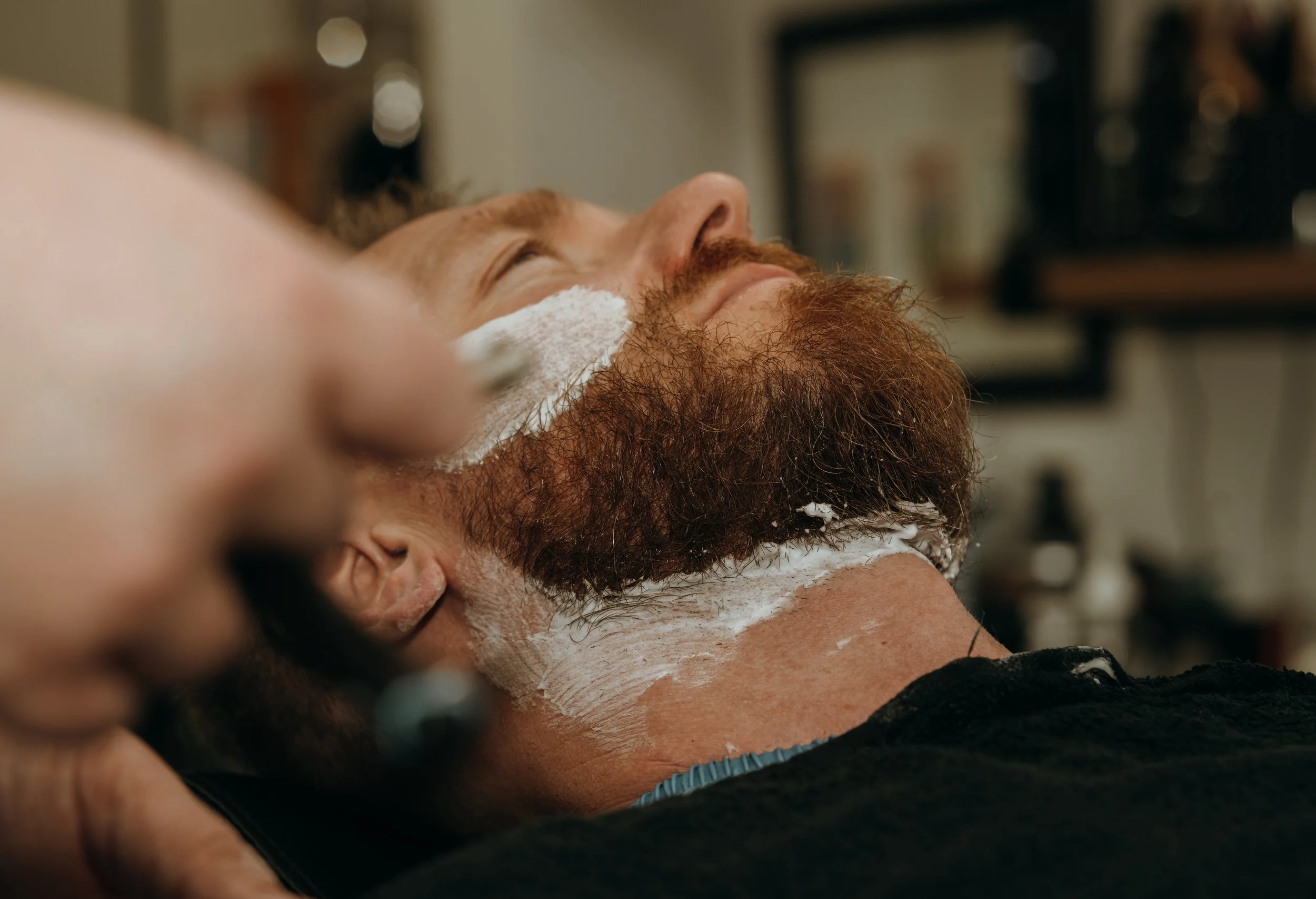 Man lying back with eyes closed while getting a shave, with shaving cream on his face and neck, in a barbershop setting.
